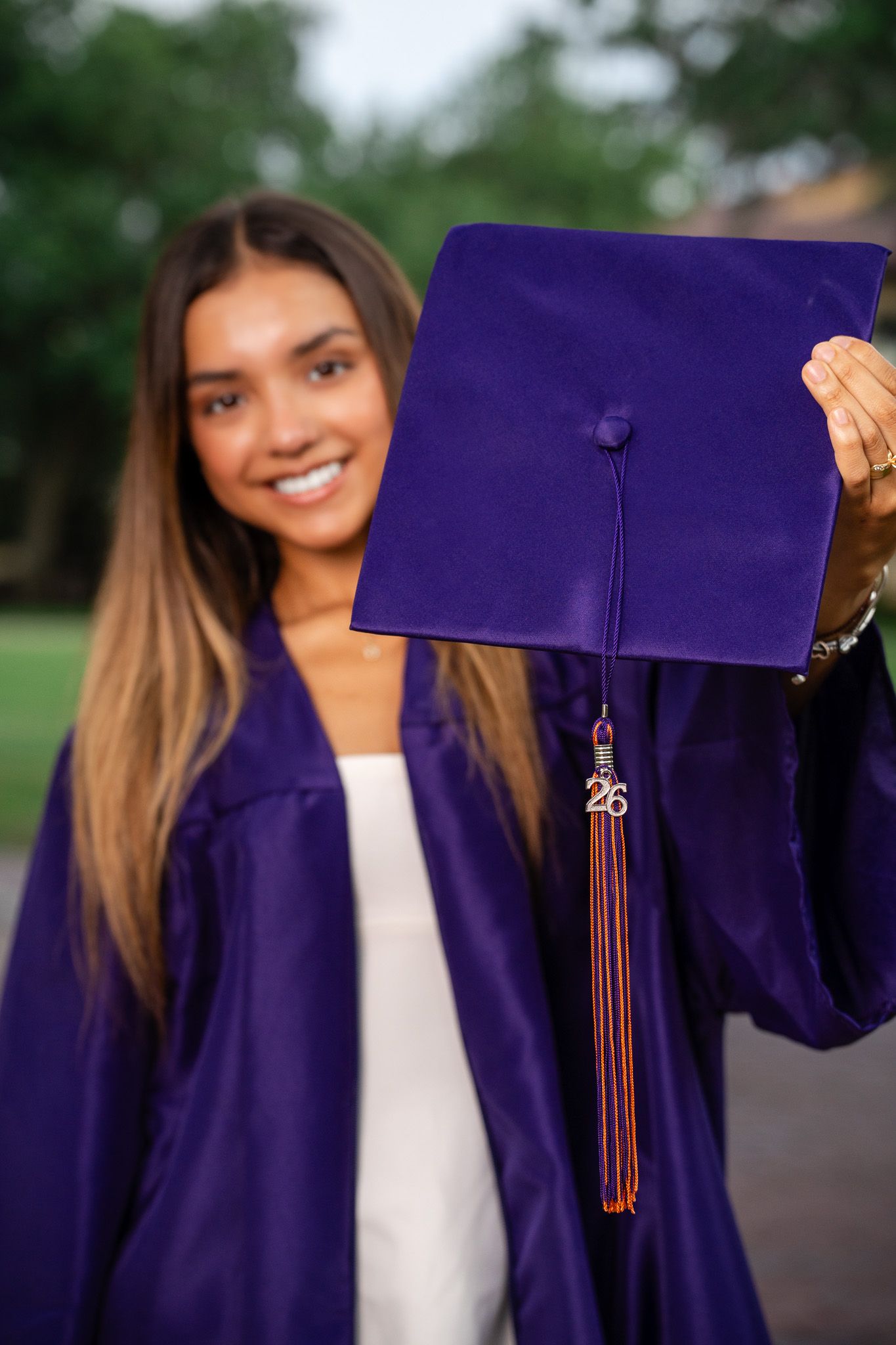 Female graduate in purple gown holds up graduation cap with tassel and charm close to camera.