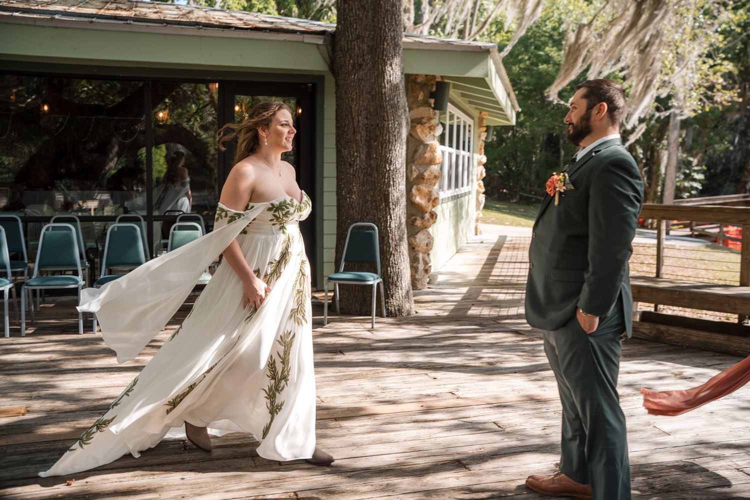 Bride in floral wedding gown twirls as groom in green suit watches during outdoor first look.