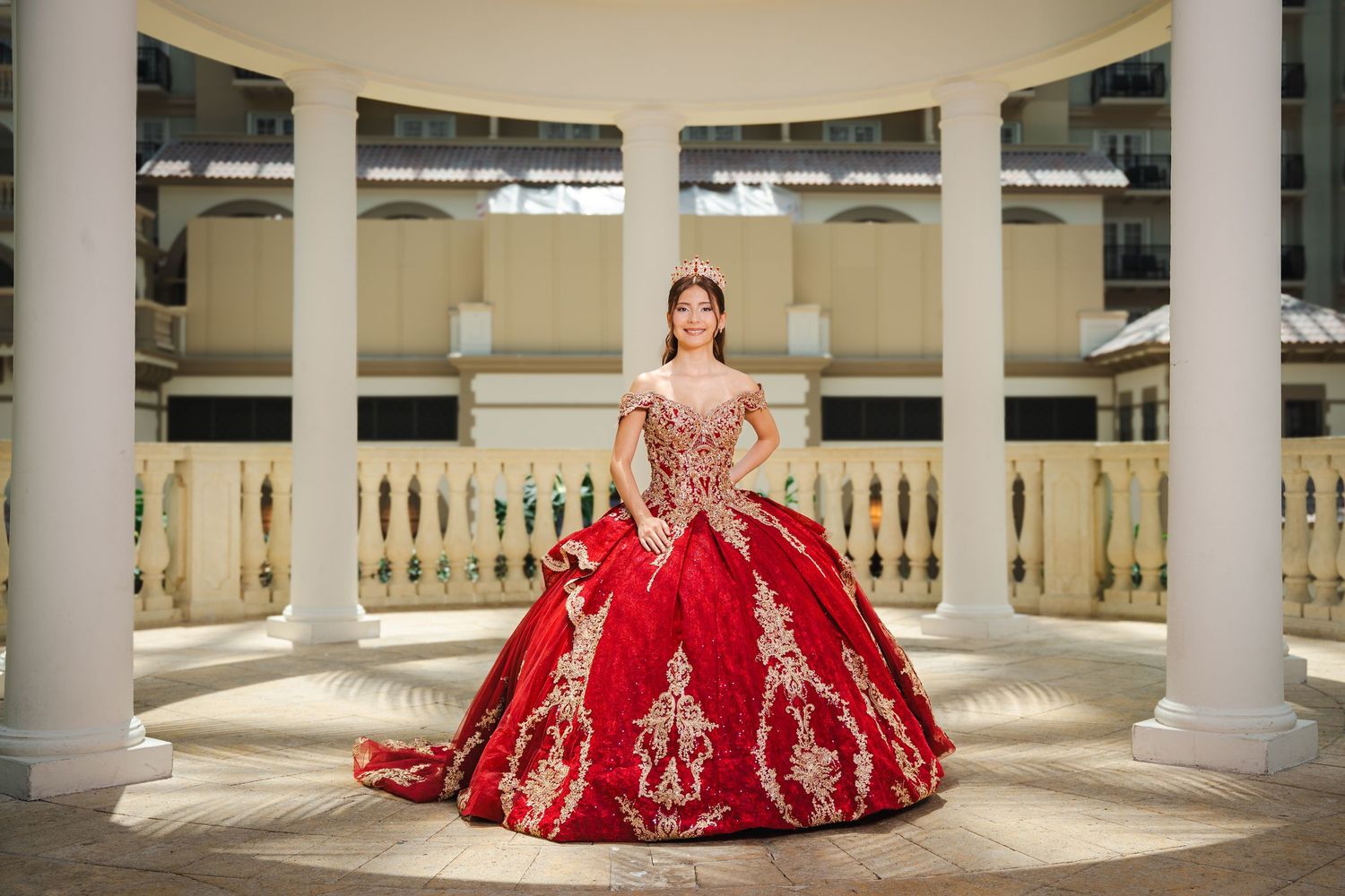 Young woman in red and gold ball gown quinceañera dress posing between white columns.