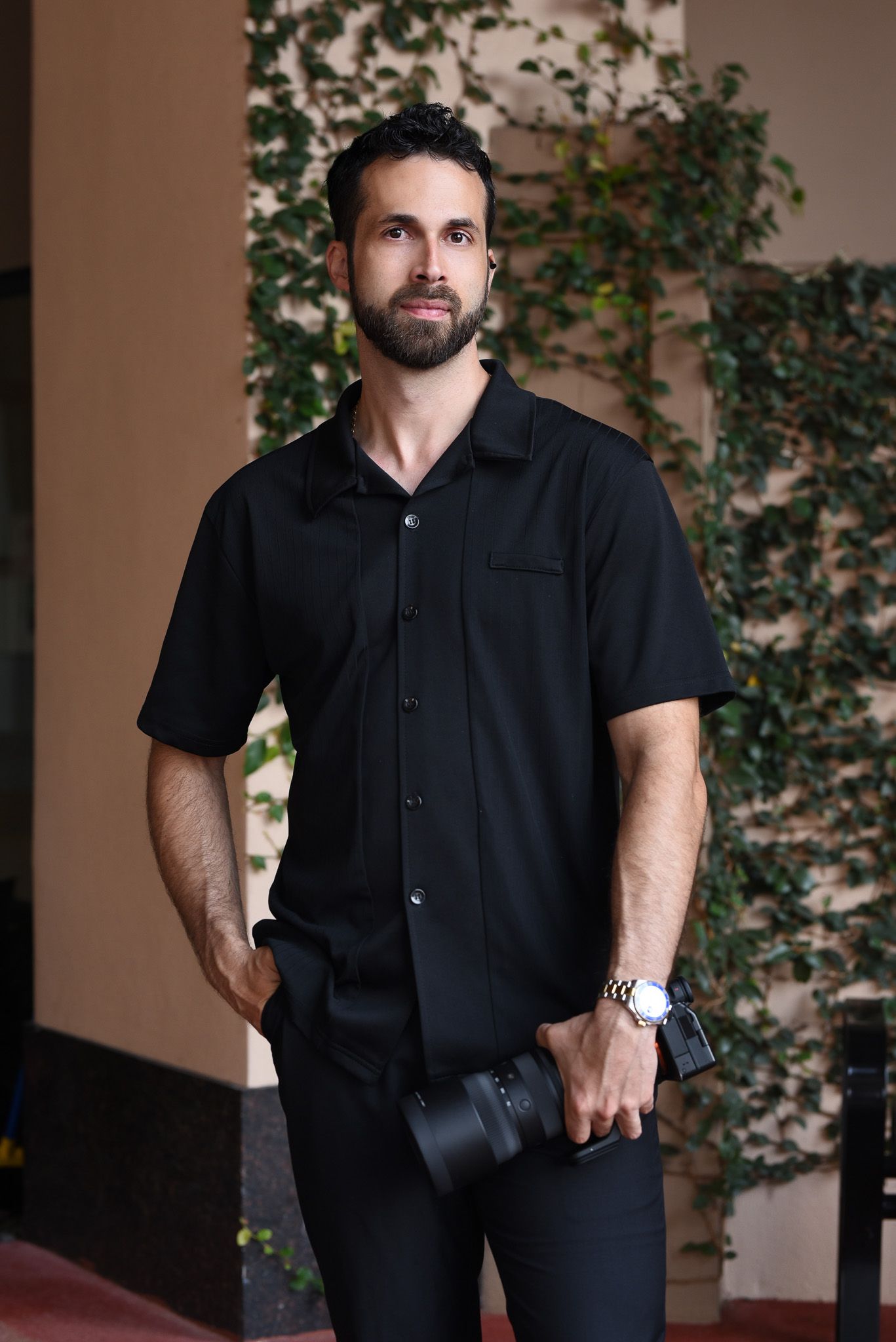 Man in black shirt holding camera with watch, standing outdoors near green ivy wall, portrait of photographer.