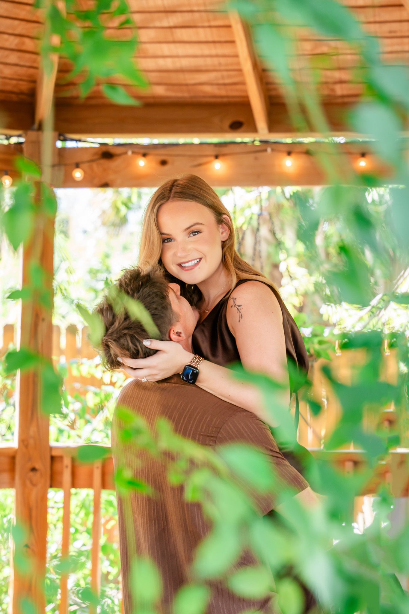 A smiling blonde woman embraces a man in a wooden gazebo surrounded by lush greenery and string lights.