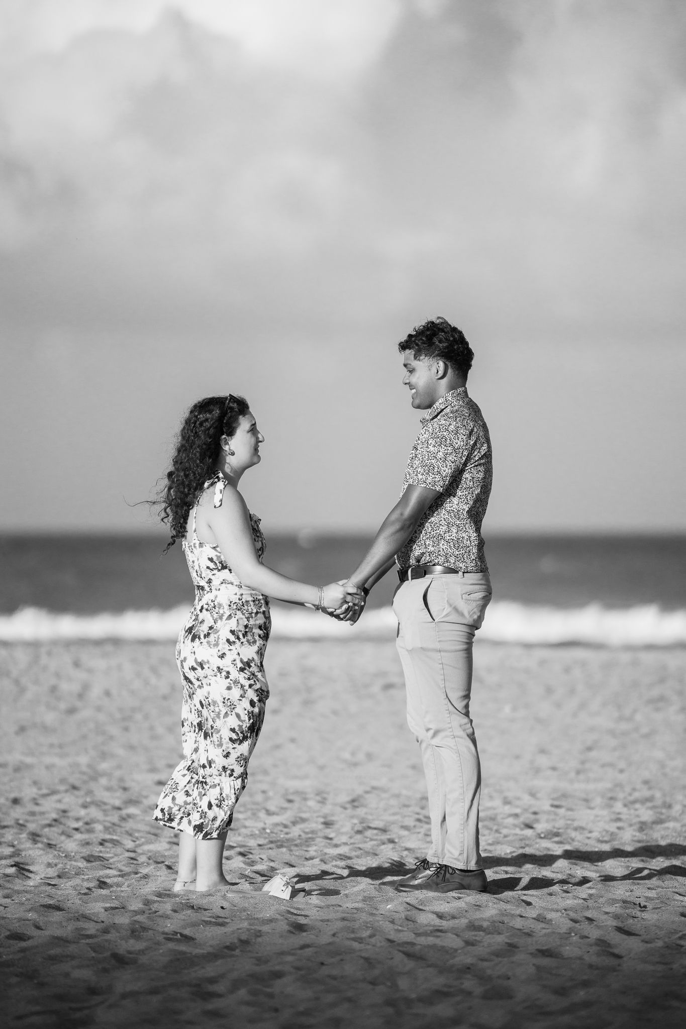 Black and white photo of a happy couple holding hands facing each other on a sandy beach with ocean waves behind them.
