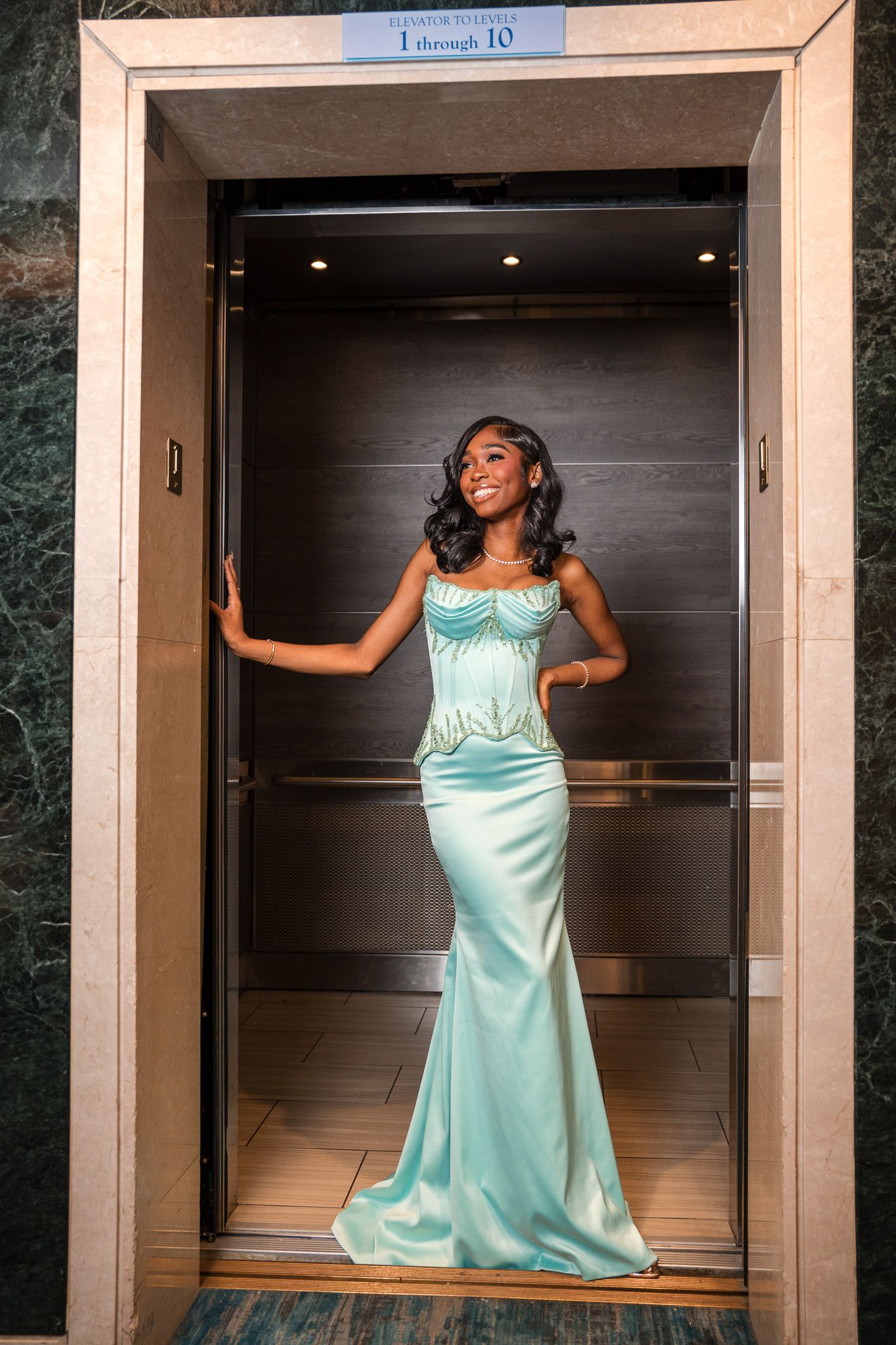 A woman in a mint green satin mermaid gown poses elegantly inside an elevator doorway.