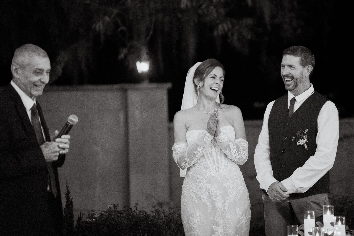 Bride and groom laugh joyfully during a wedding toast speech at their outdoor evening reception.
