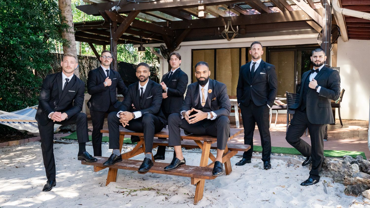 Groom and groomsmen in black suits pose confidently near a wooden bench in an outdoor setting.