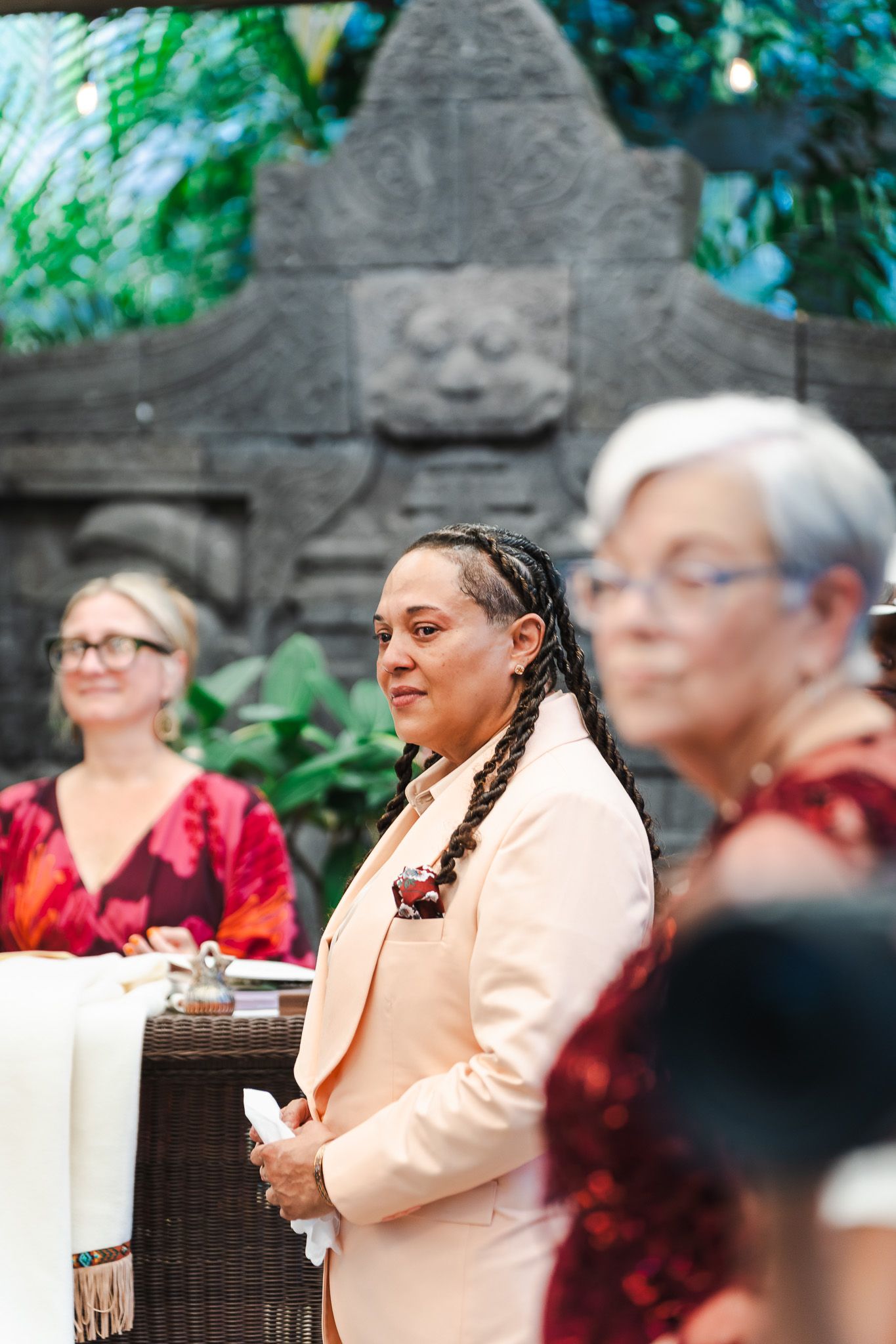 Person in peach blazer with braided hair standing attentively during an outdoor garden wedding ceremony.