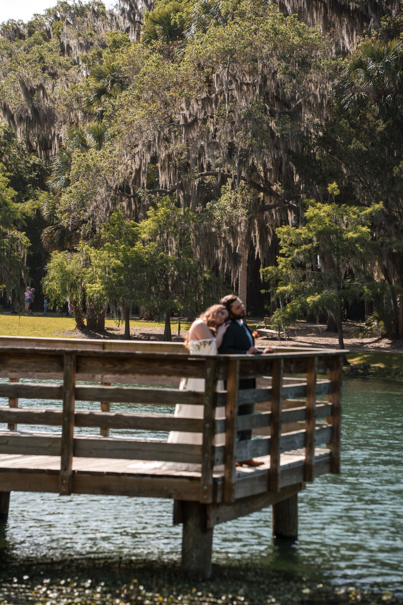 Bride and groom share a romantic moment on a wooden dock overlooking a serene lake surrounded by moss-draped trees.