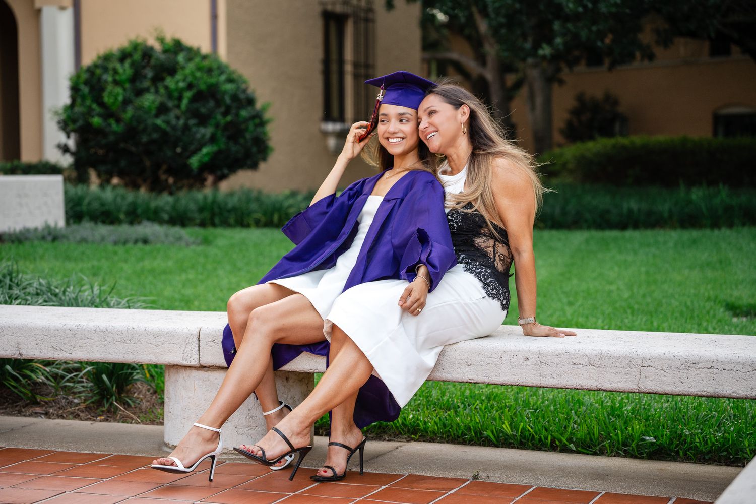 Graduate in purple cap and gown sitting with proud mother on outdoor ledge, sharing a joyful moment.