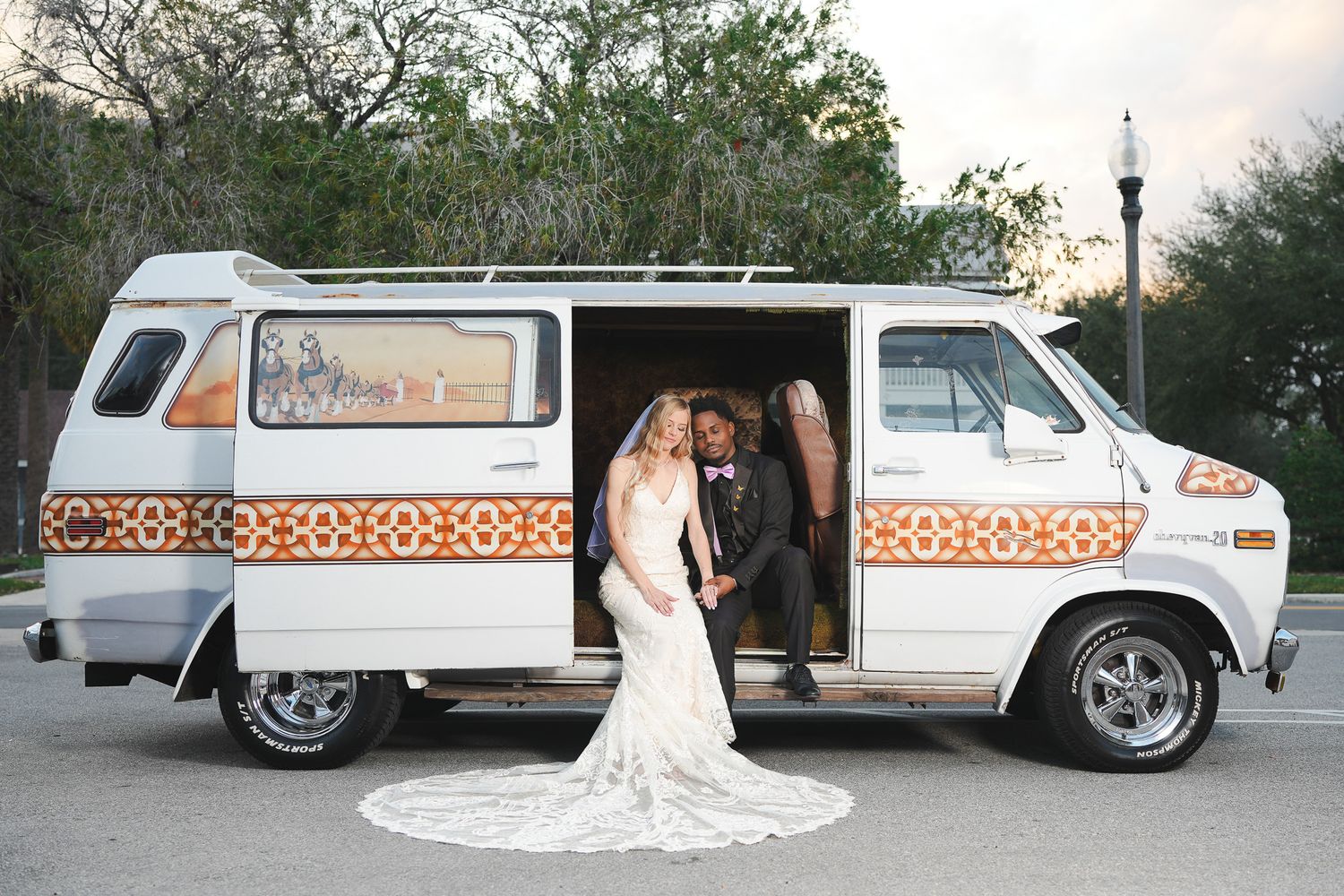 Bride and groom pose together in front of a vintage white decorated van at their outdoor wedding.