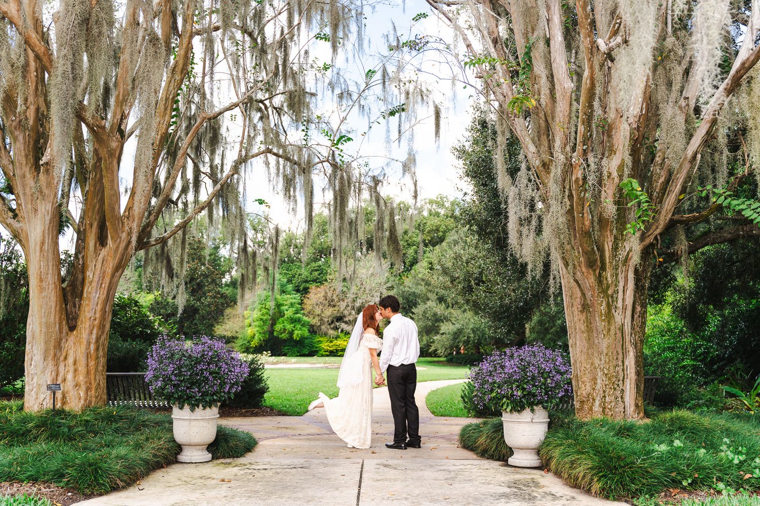 Bride and groom share a romantic moment on a garden pathway lined with moss-draped trees and purple flowering plants.