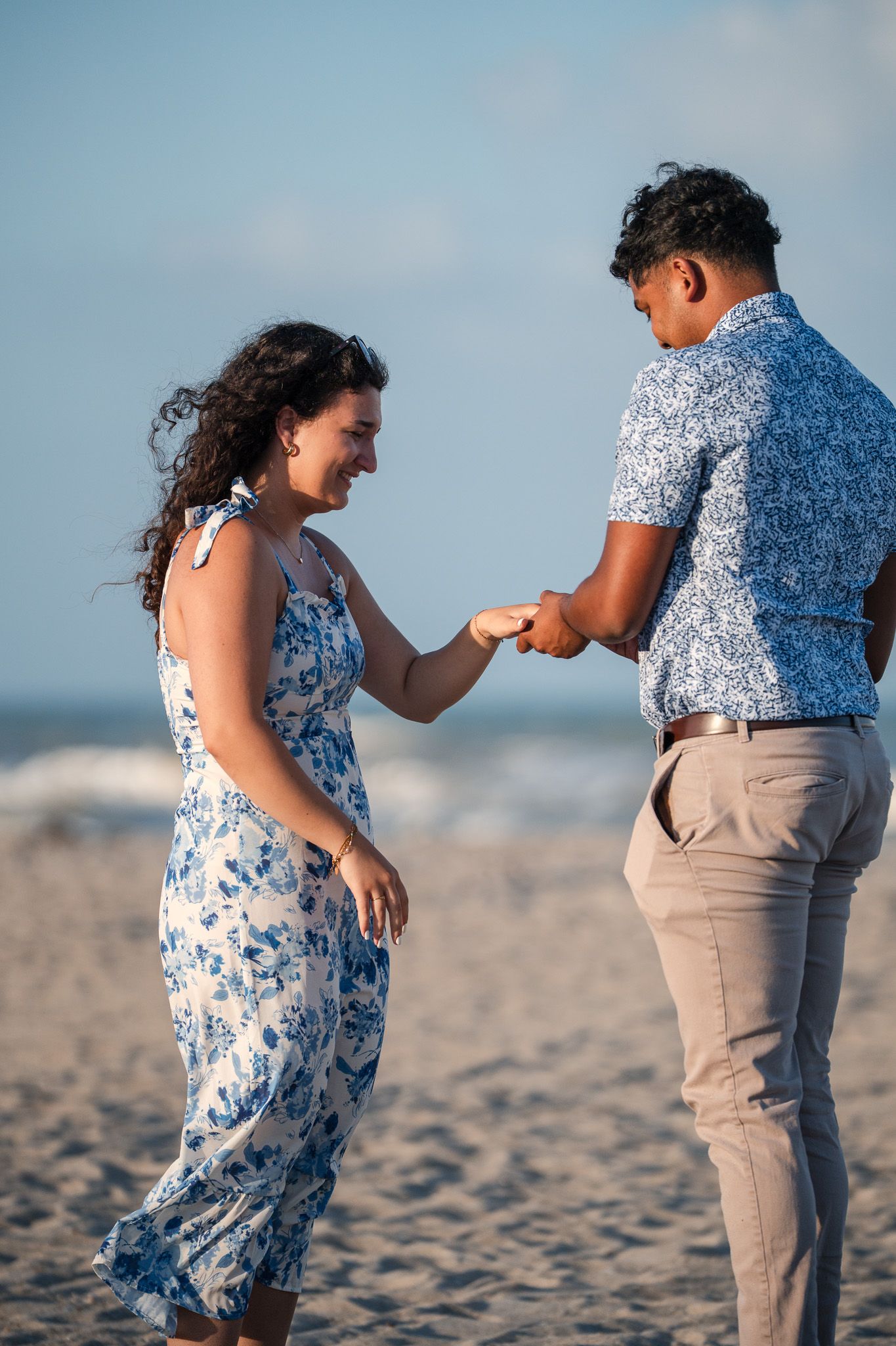 A couple in matching blue floral outfits share a romantic moment on a sandy beach, as the man places a ring on her finger.