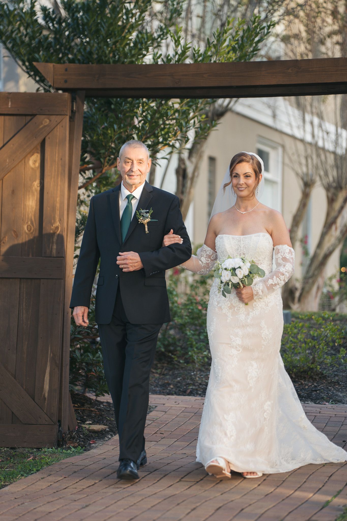Father proudly walks bride in lace gown down brick path through wooden garden gate.