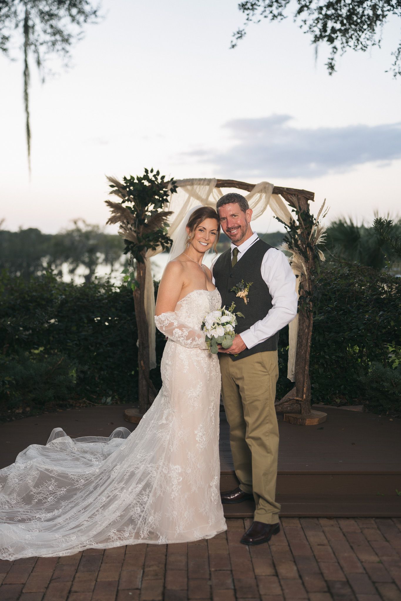 Smiling bride and groom pose together before rustic wooden arch at waterfront wedding.
