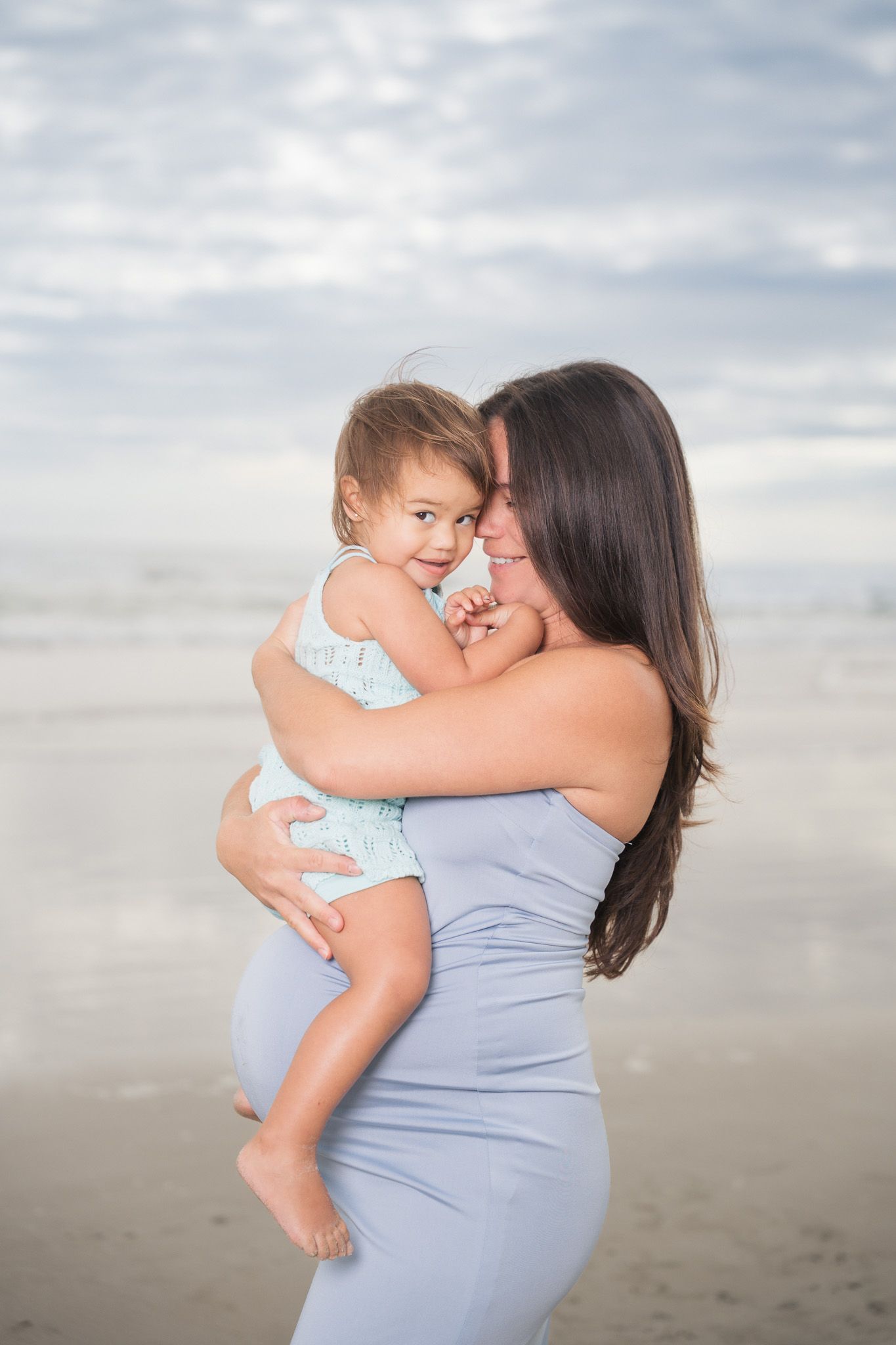 Pregnant woman in blue dress tenderly holds and gazes at smiling toddler on beach.