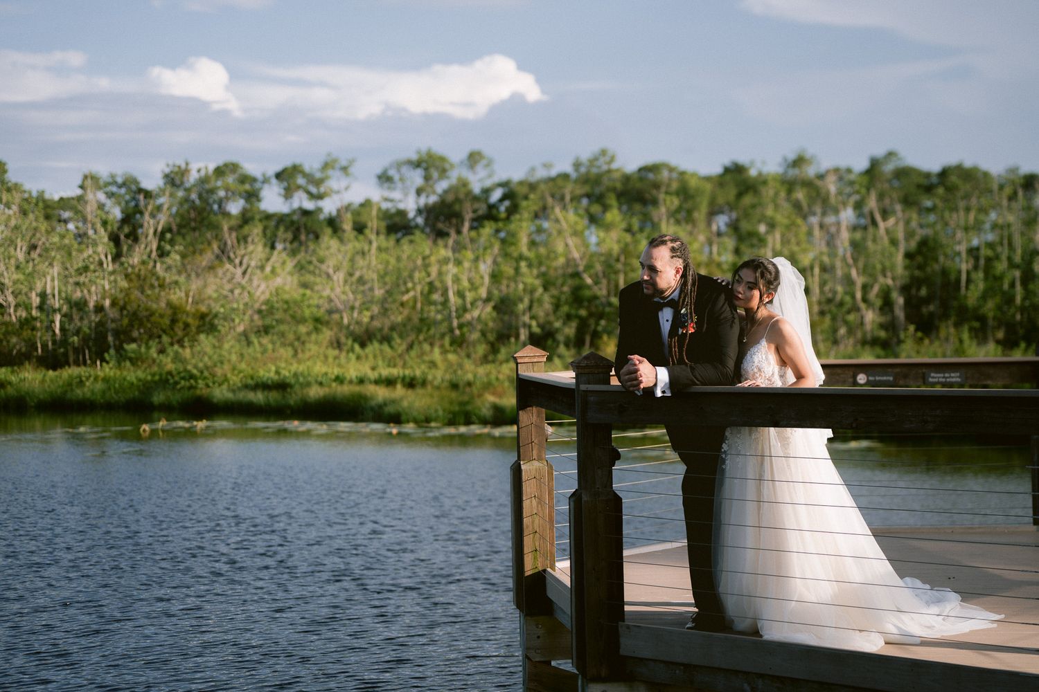 Bride and groom share romantic moment on wooden dock overlooking serene lake with lush greenery.