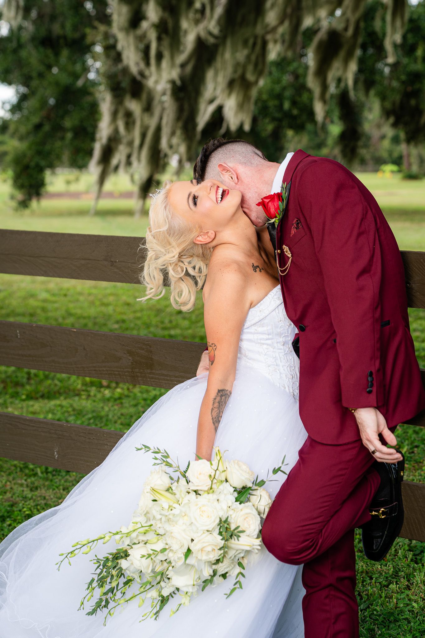 Groom dips laughing bride in white strapless gown holding white bouquet against rustic wooden fence outdoors.