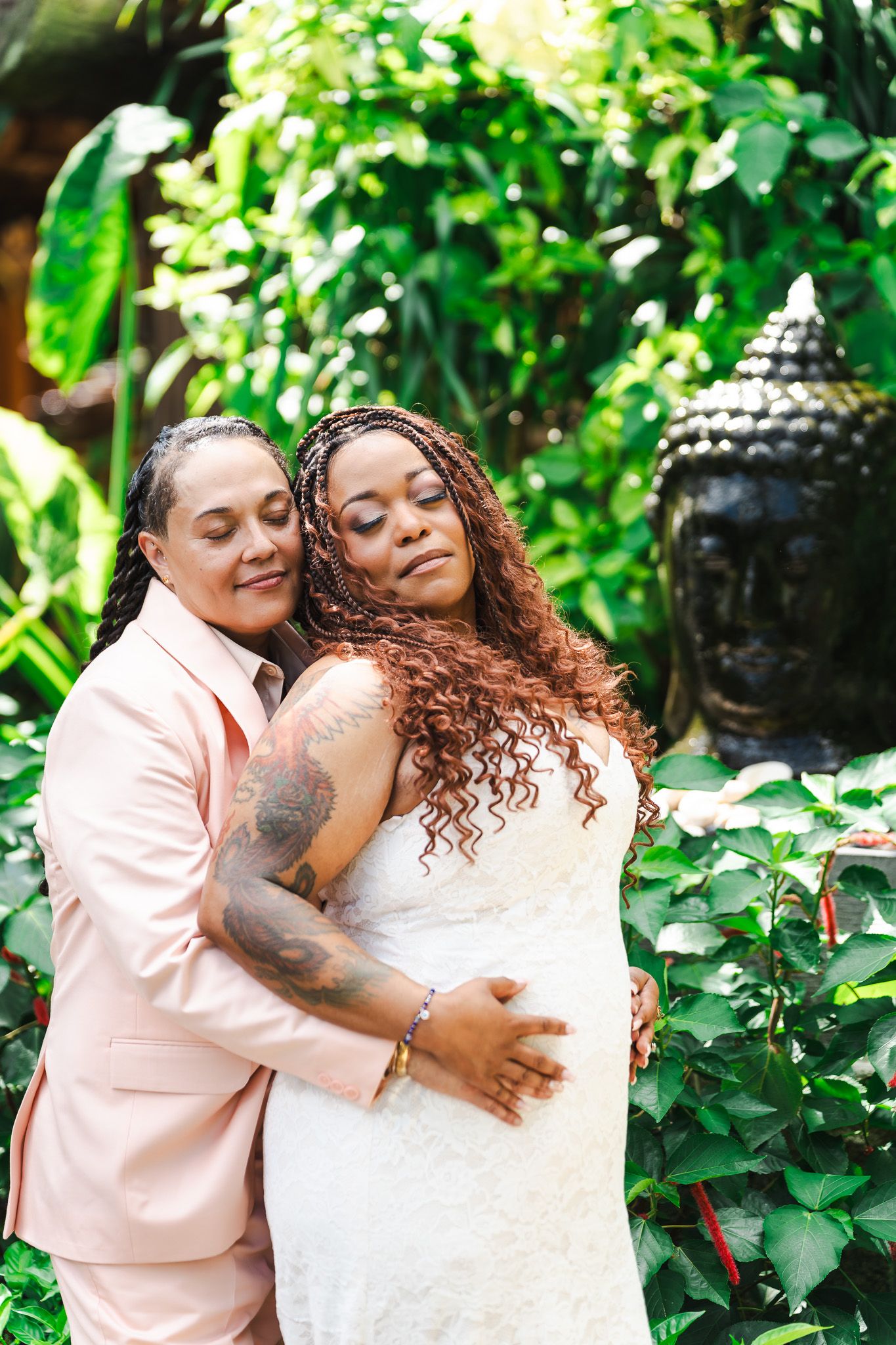 Two brides share tender embrace in lush garden with Buddha statue backdrop at wedding.