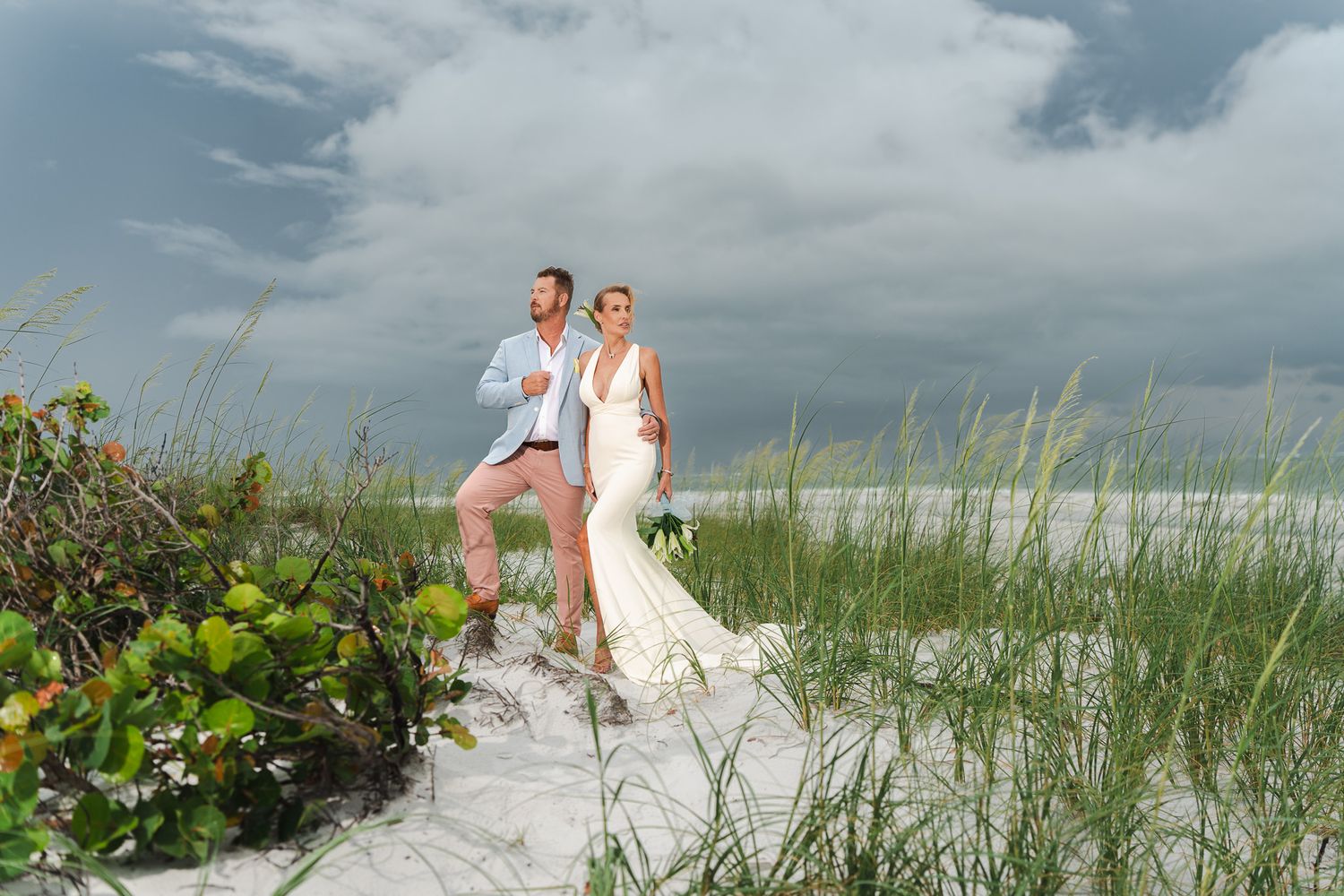 Bride and groom standing together on white sand beach dunes with sea grass and stormy clouds behind them.