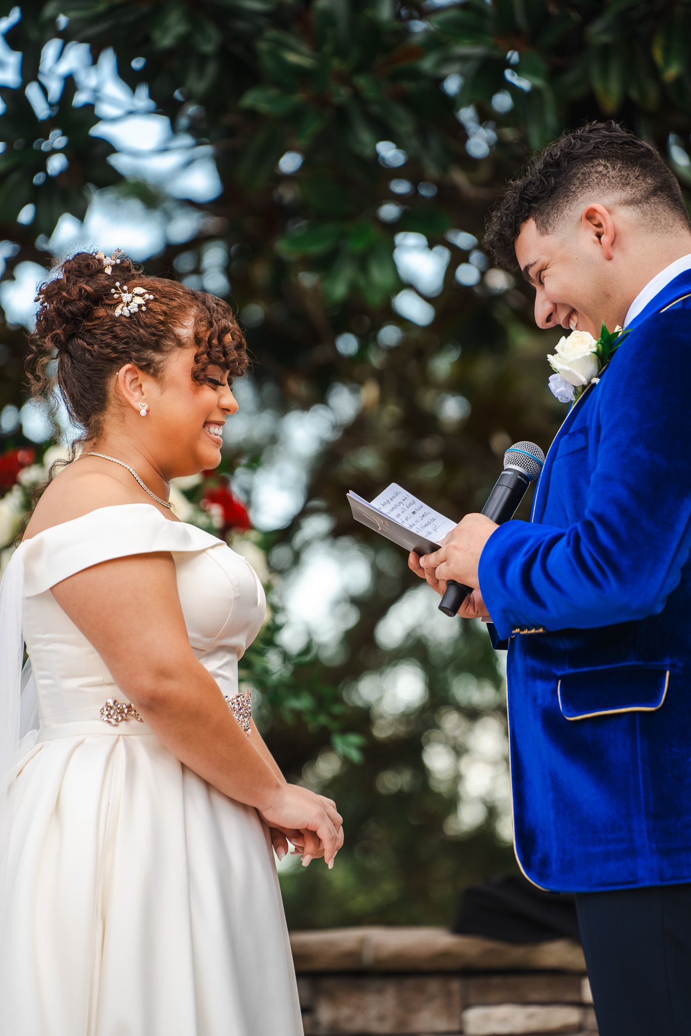 Groom in blue velvet jacket laughs while reading vows to smiling bride at outdoor ceremony.
