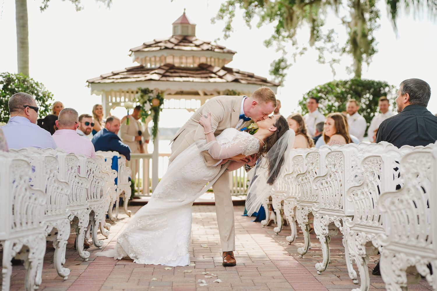 Groom dips bride for a kiss at outdoor gazebo wedding ceremony with white ornate chairs and guests.