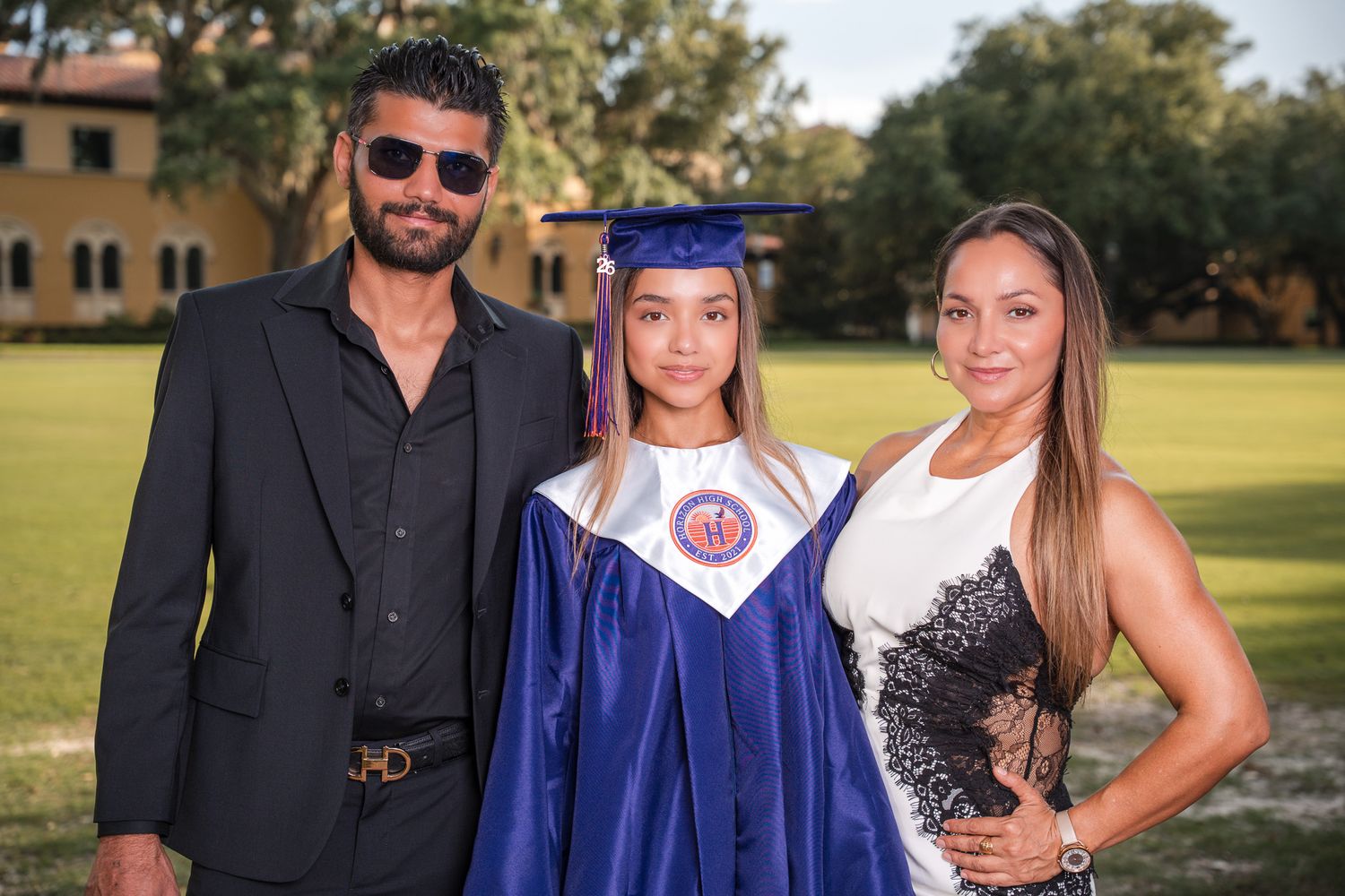 Graduate in cap and gown poses with proud parents outdoors on a sunny day at a college campus.