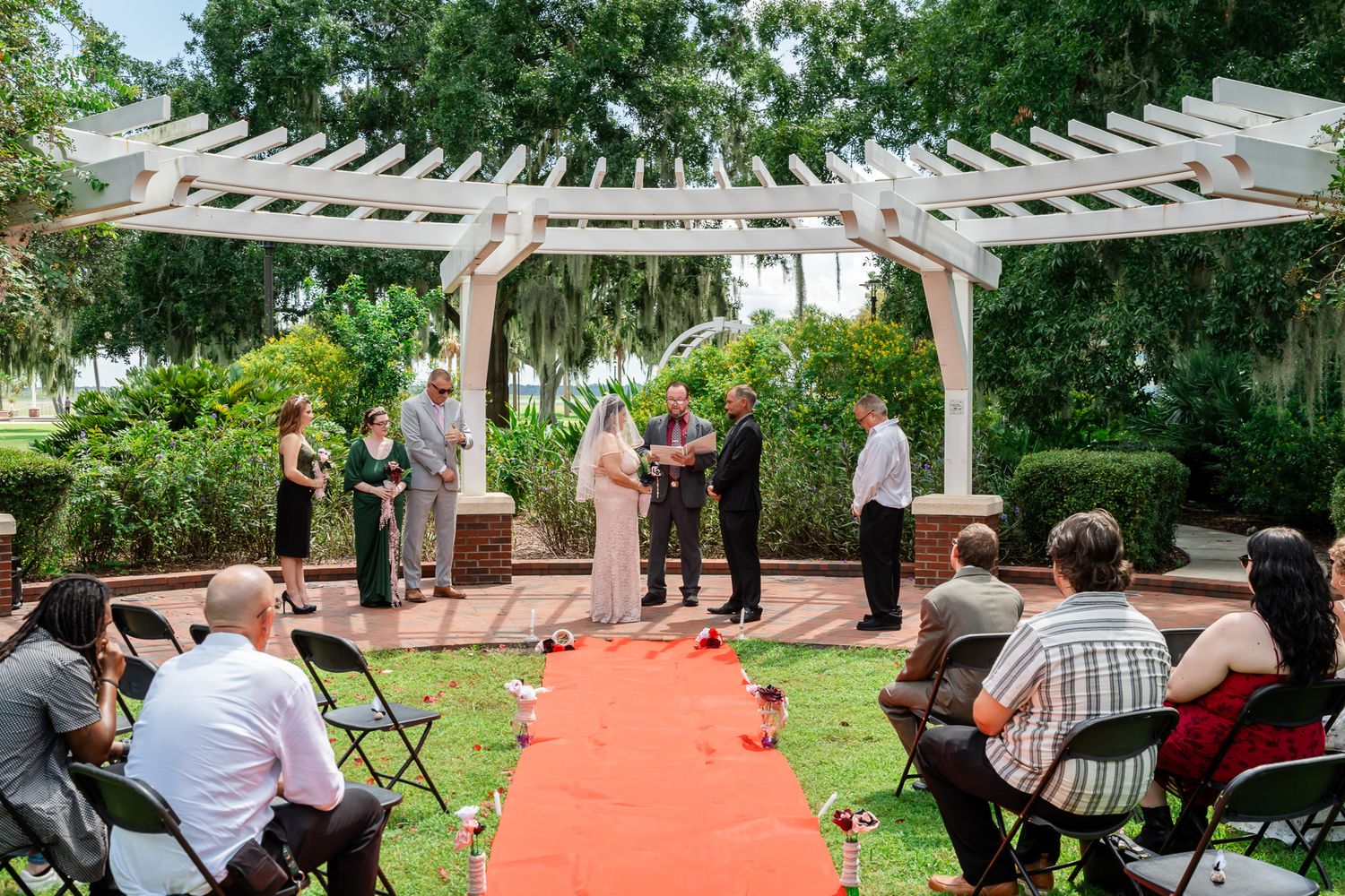 Outdoor wedding ceremony under white pergola with red carpet aisle, guests seated on black chairs surrounded by lush greenery.