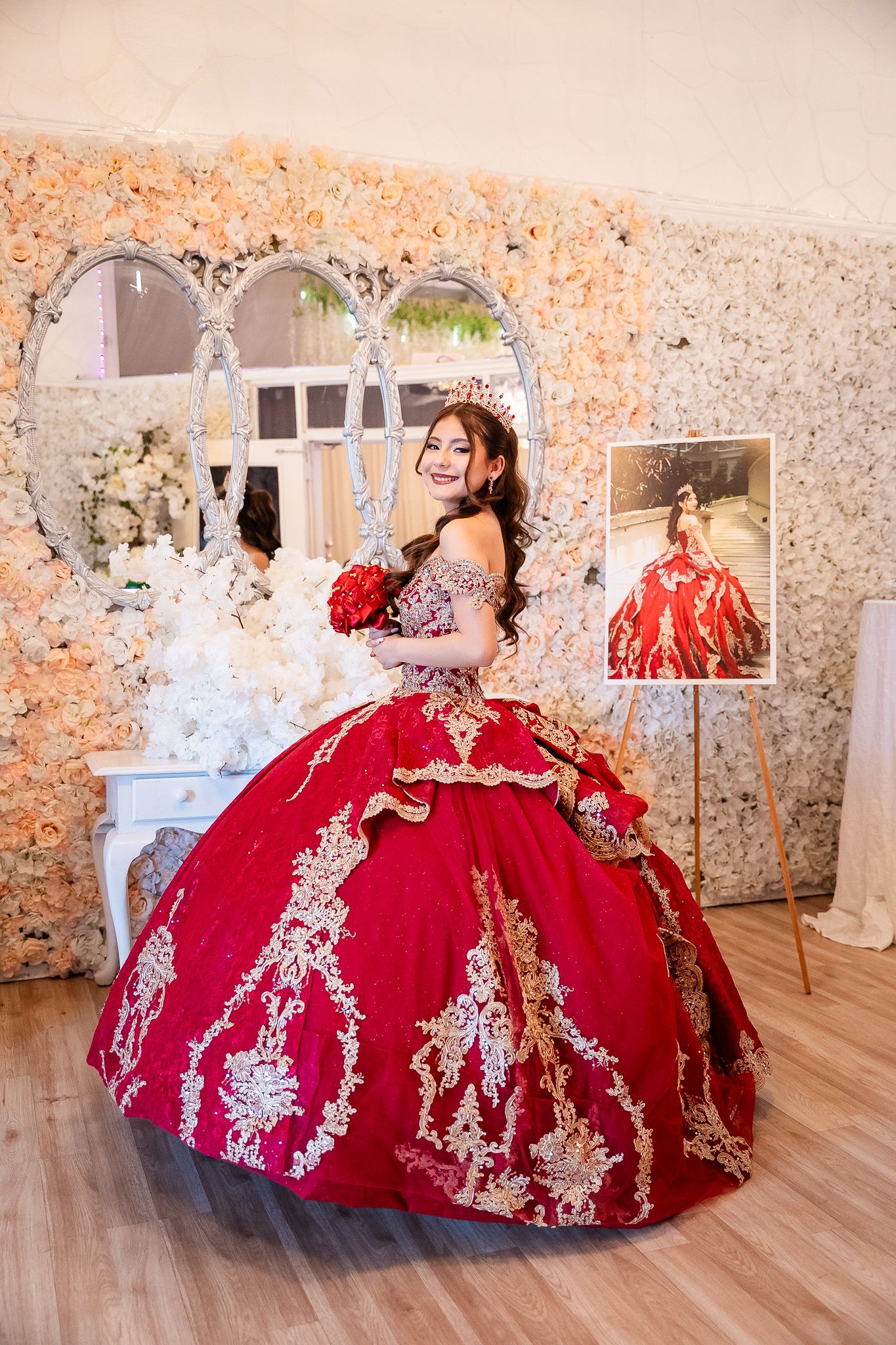 Young woman in ornate red and gold quinceañera ball gown poses by floral wall with mirror.