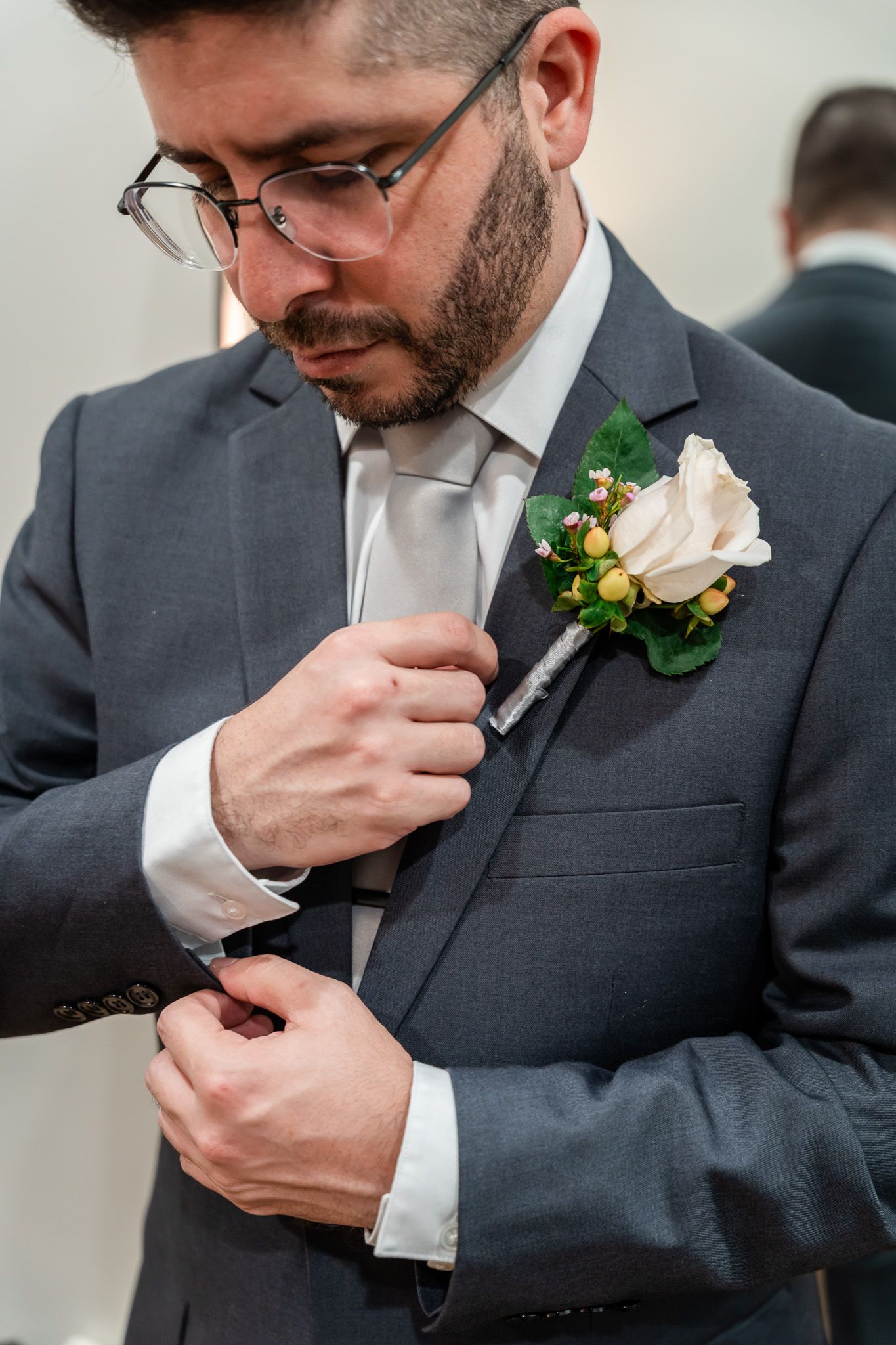Dapper groom in gray suit adjusting cufflinks with white rose boutonniere on lapel.