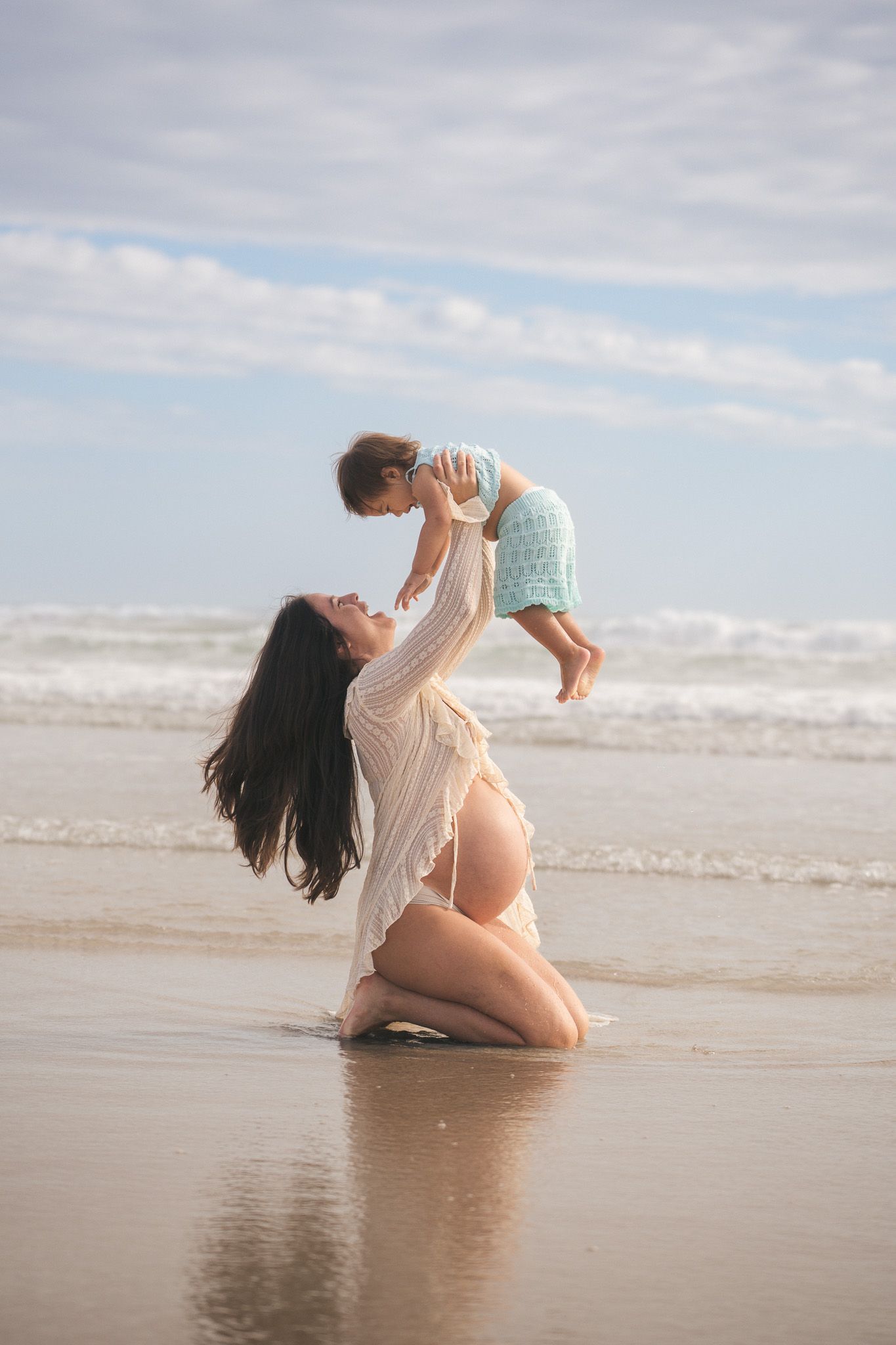 Pregnant mother kneels on wet sand lifting her baby joyfully during a golden hour beach maternity session.