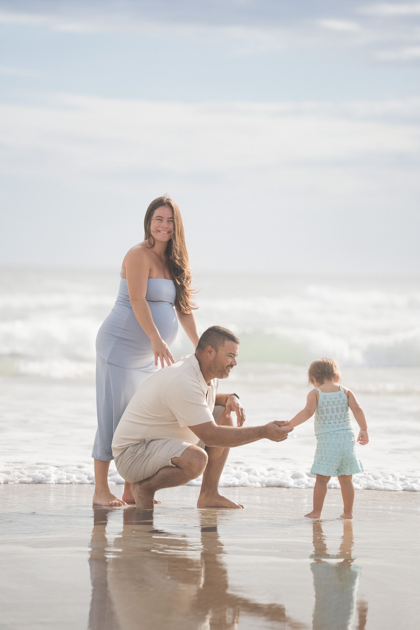 Family of three enjoys playful moment at ocean shoreline with pregnant mother and toddler.