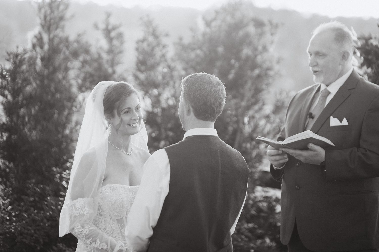 Smiling bride exchanges vows with groom before officiant in black and white ceremony photo.