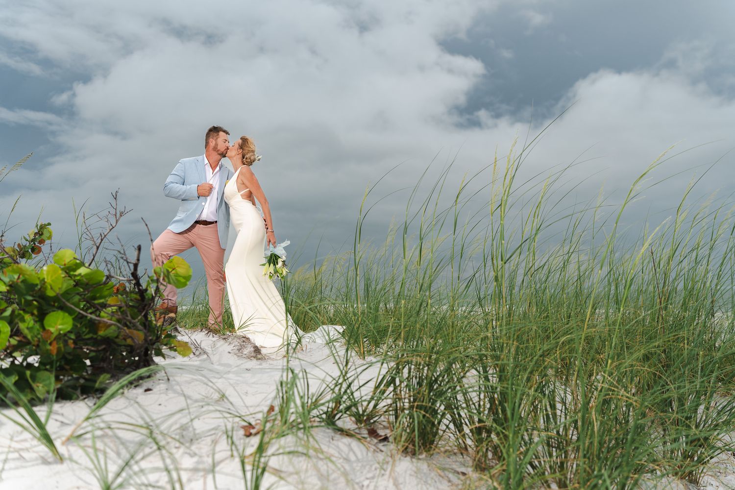 Bride and groom sharing romantic kiss on white sand dunes surrounded by sea grass at beach wedding.