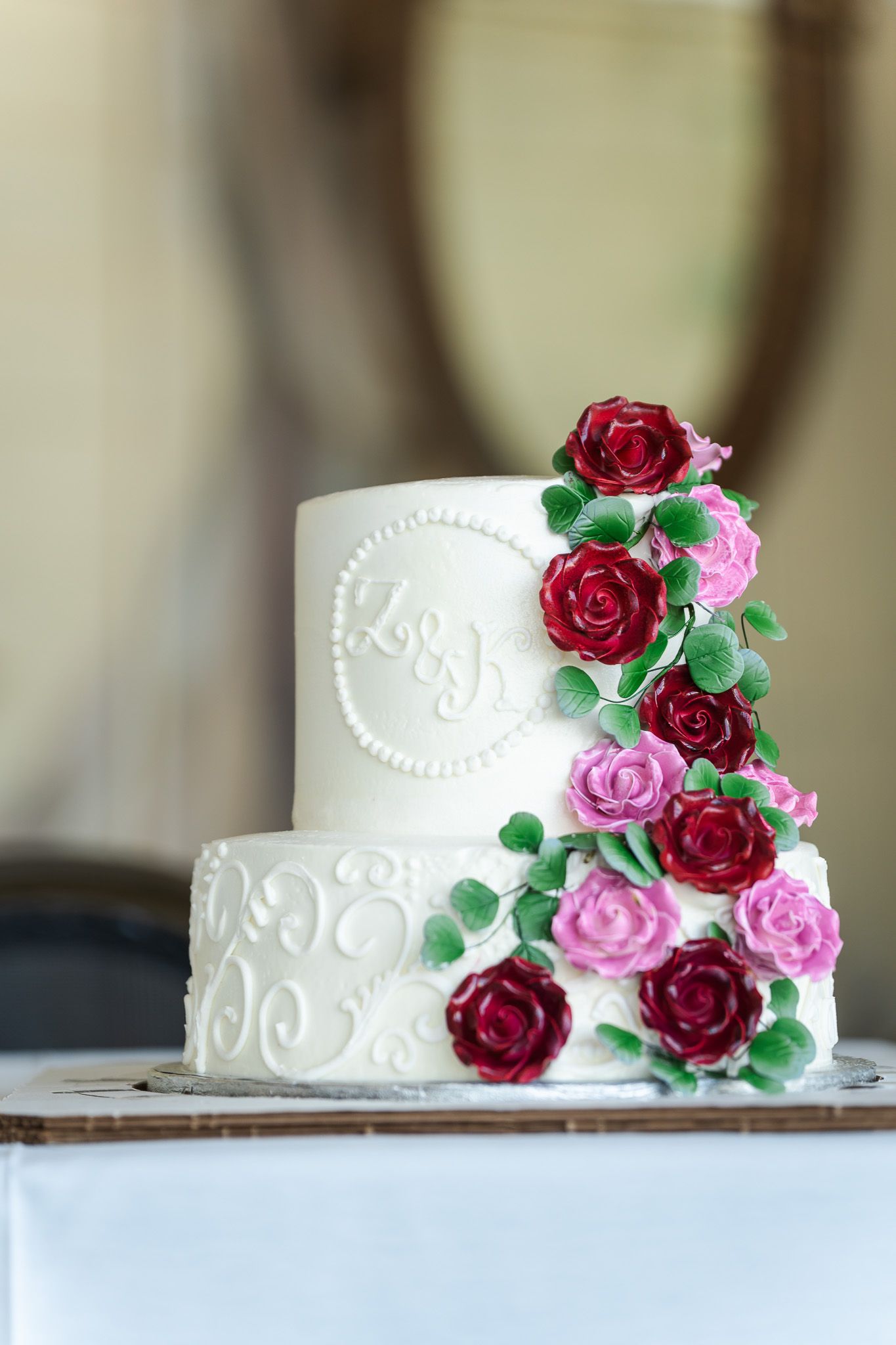 Elegant two-tier white wedding cake decorated with red and pink sugar roses and green leaves.