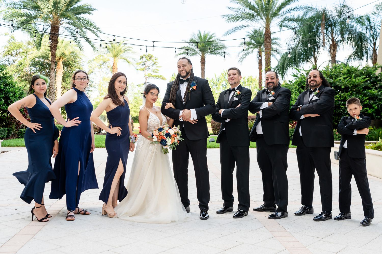 Wedding party in navy and black formal attire posing outdoors with palm trees.