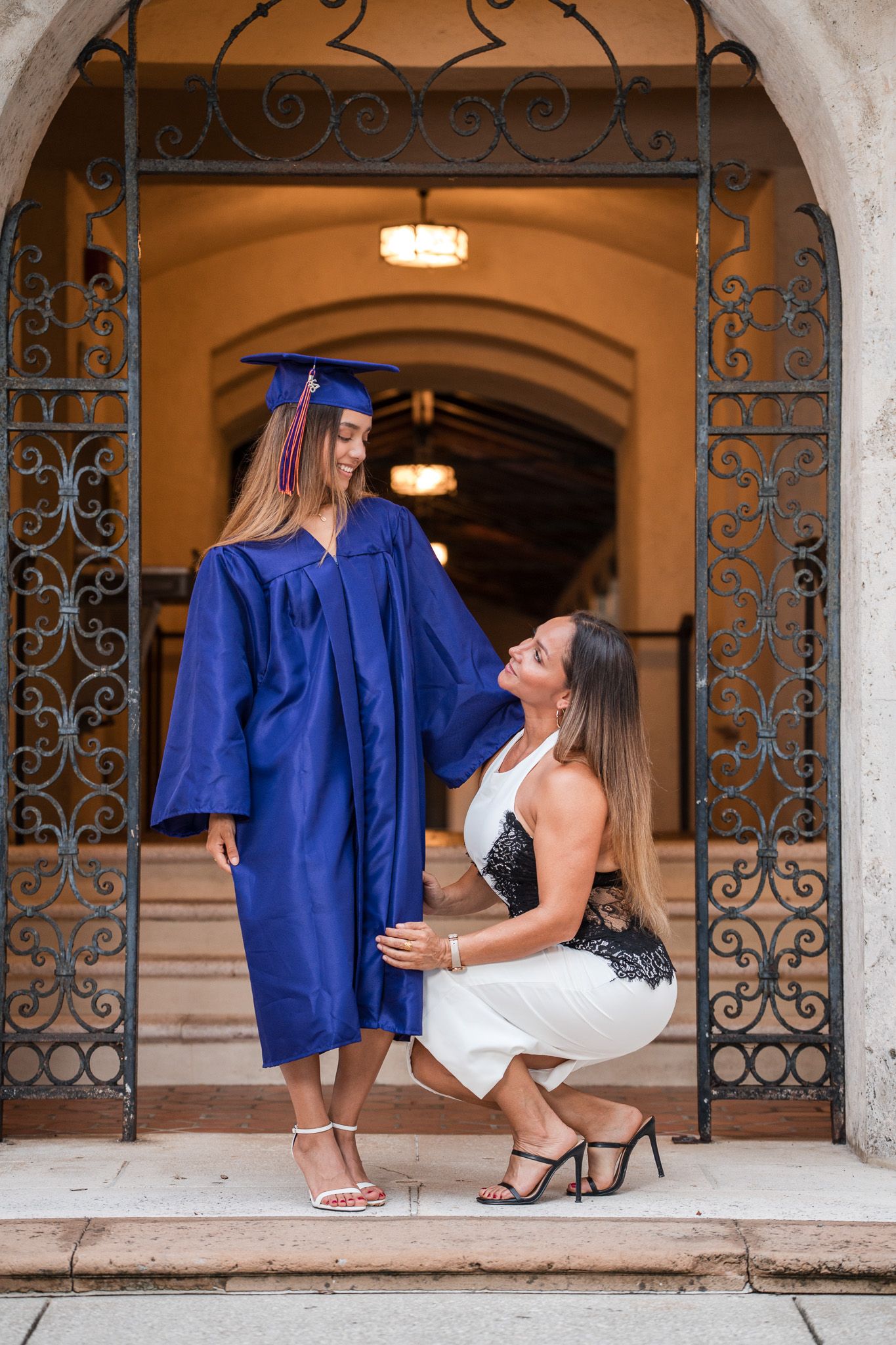 Graduate in blue cap and gown poses with proud mother in white dress at ornate iron gate entrance.