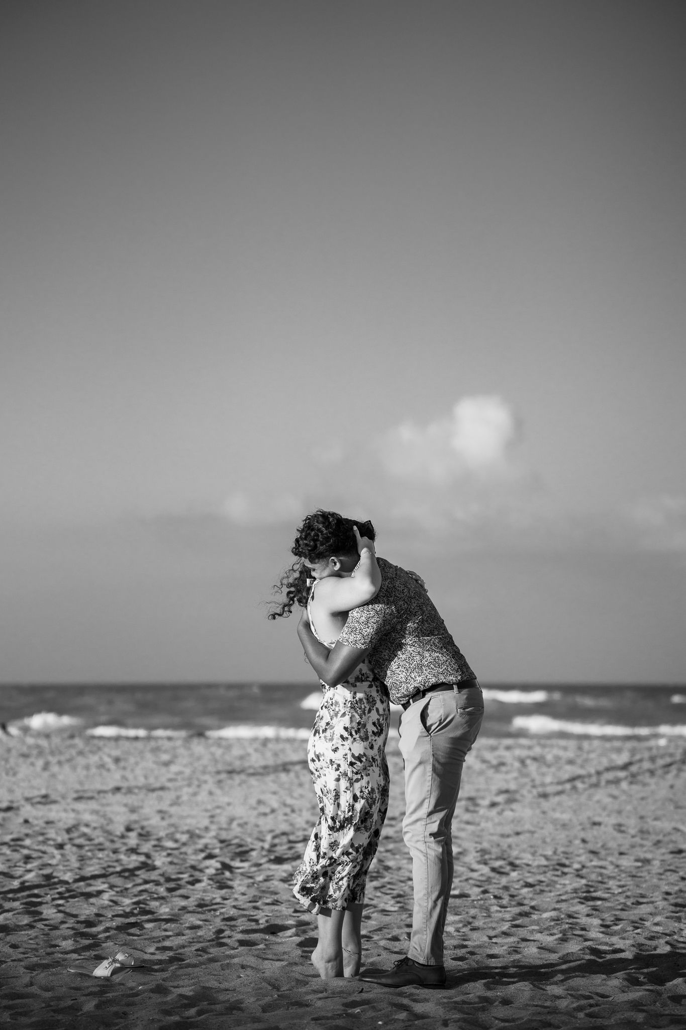 Black and white photo of a couple embracing and kissing on a sandy beach with ocean waves in the background.