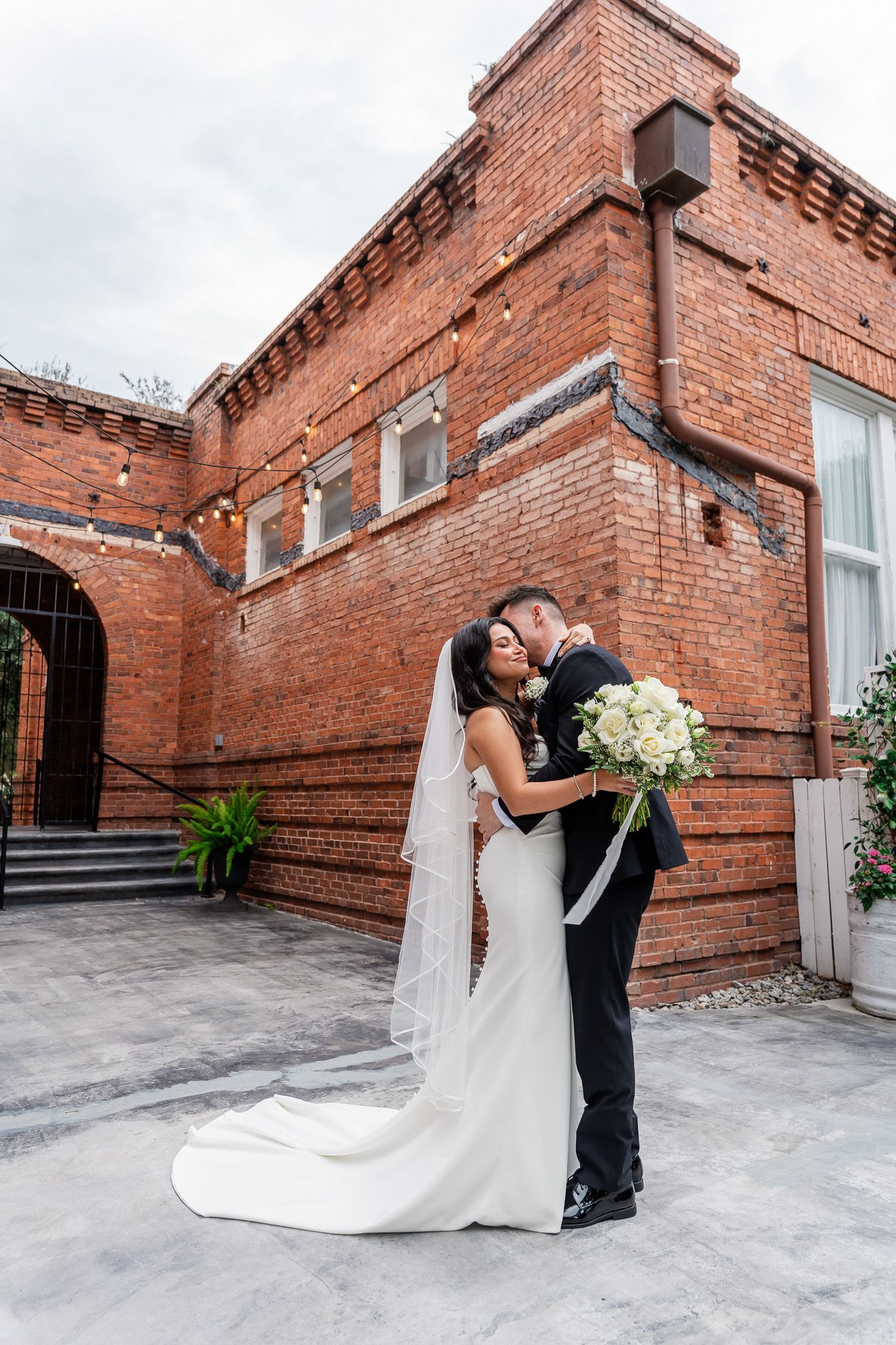 Bride and groom share a romantic embrace outside a historic red brick wedding venue.