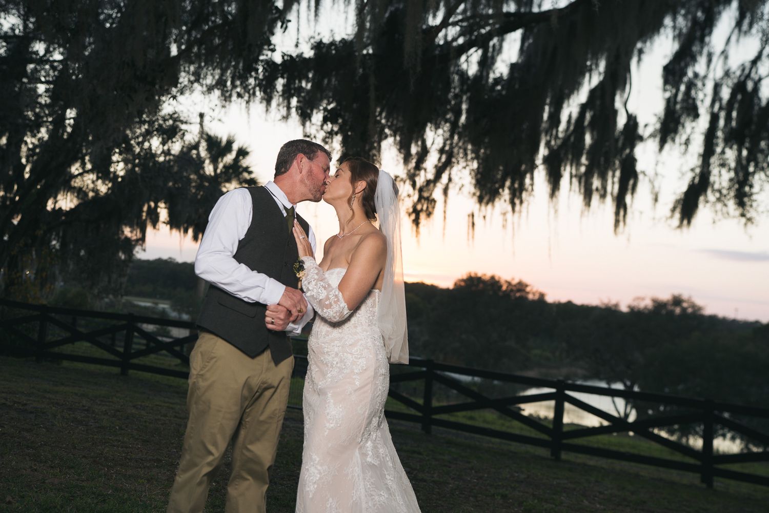 Bride and groom share romantic sunset kiss under Spanish moss-draped trees at wedding.