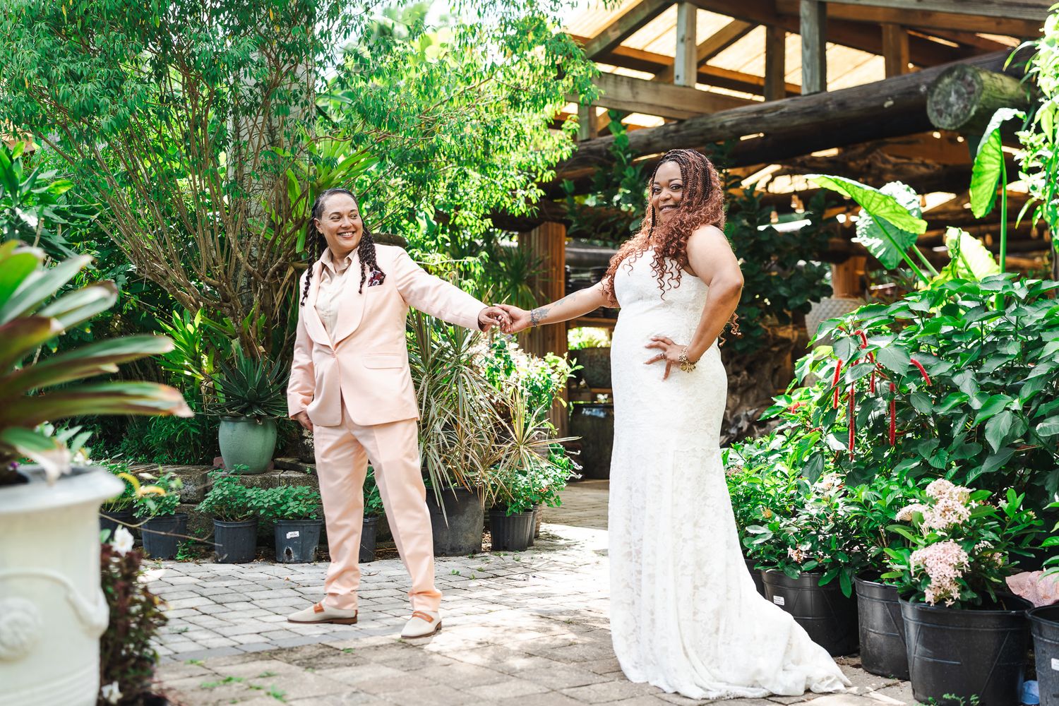 Two brides hold hands and smile in tropical garden setting during wedding celebration outdoors.