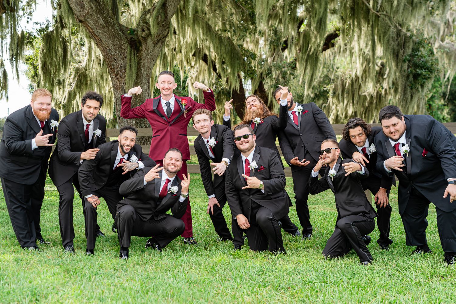 Groom in burgundy suit poses playfully with groomsmen in black suits under a Spanish moss-covered tree.