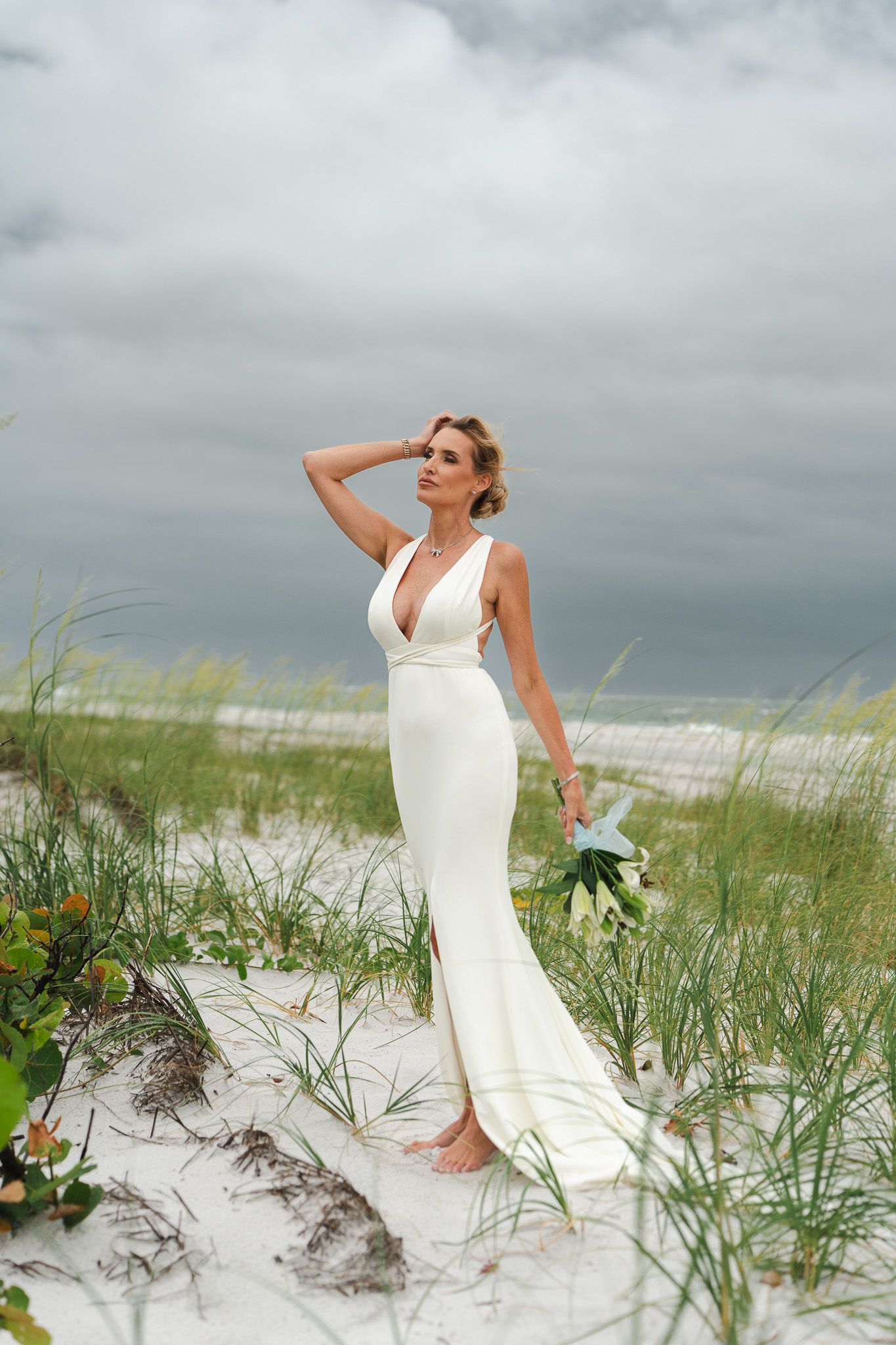 Barefoot bride in fitted white wedding gown standing on sand dunes with sea grass under dramatic cloudy sky.