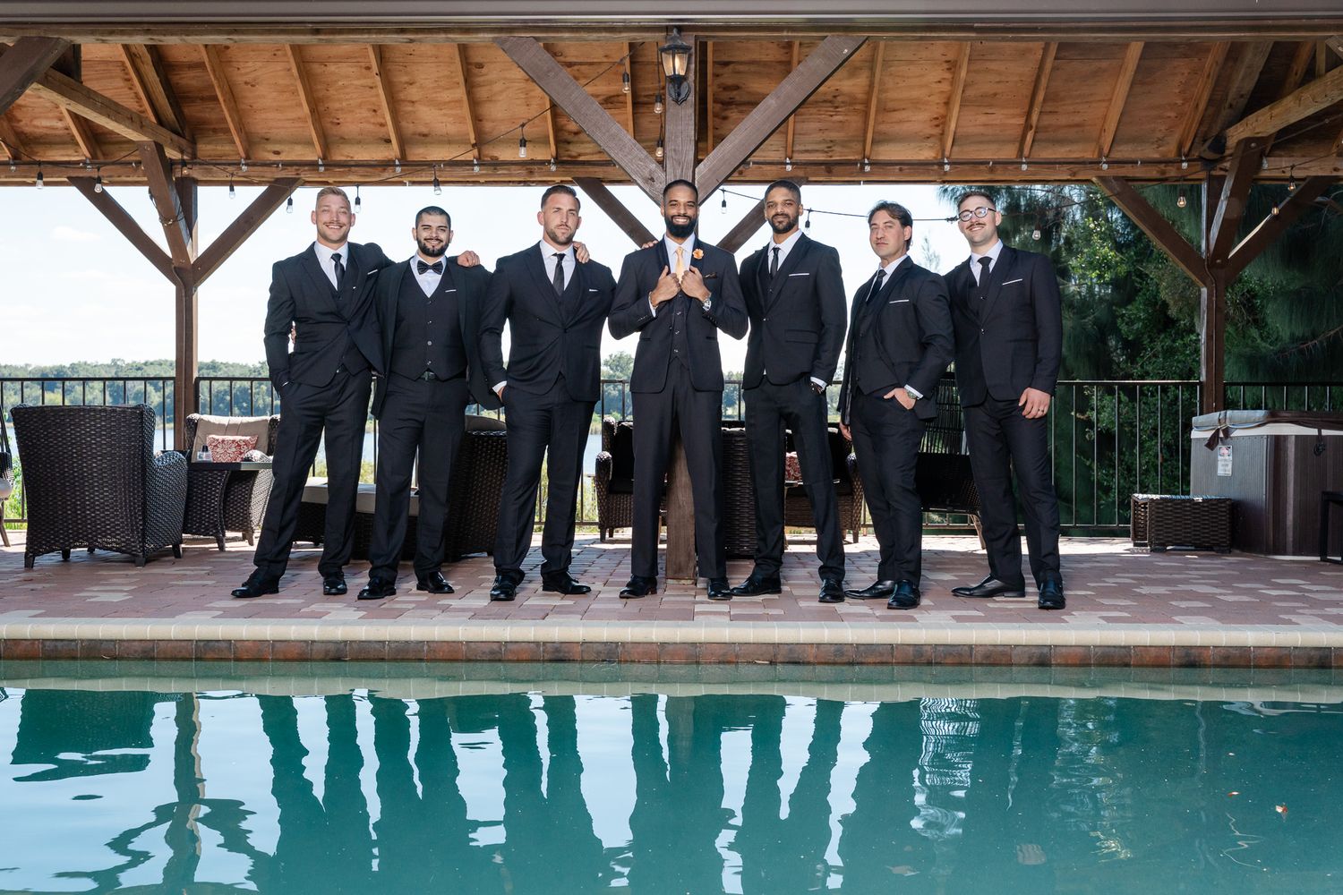 Groom and groomsmen in black suits pose confidently poolside under a wooden pavilion.