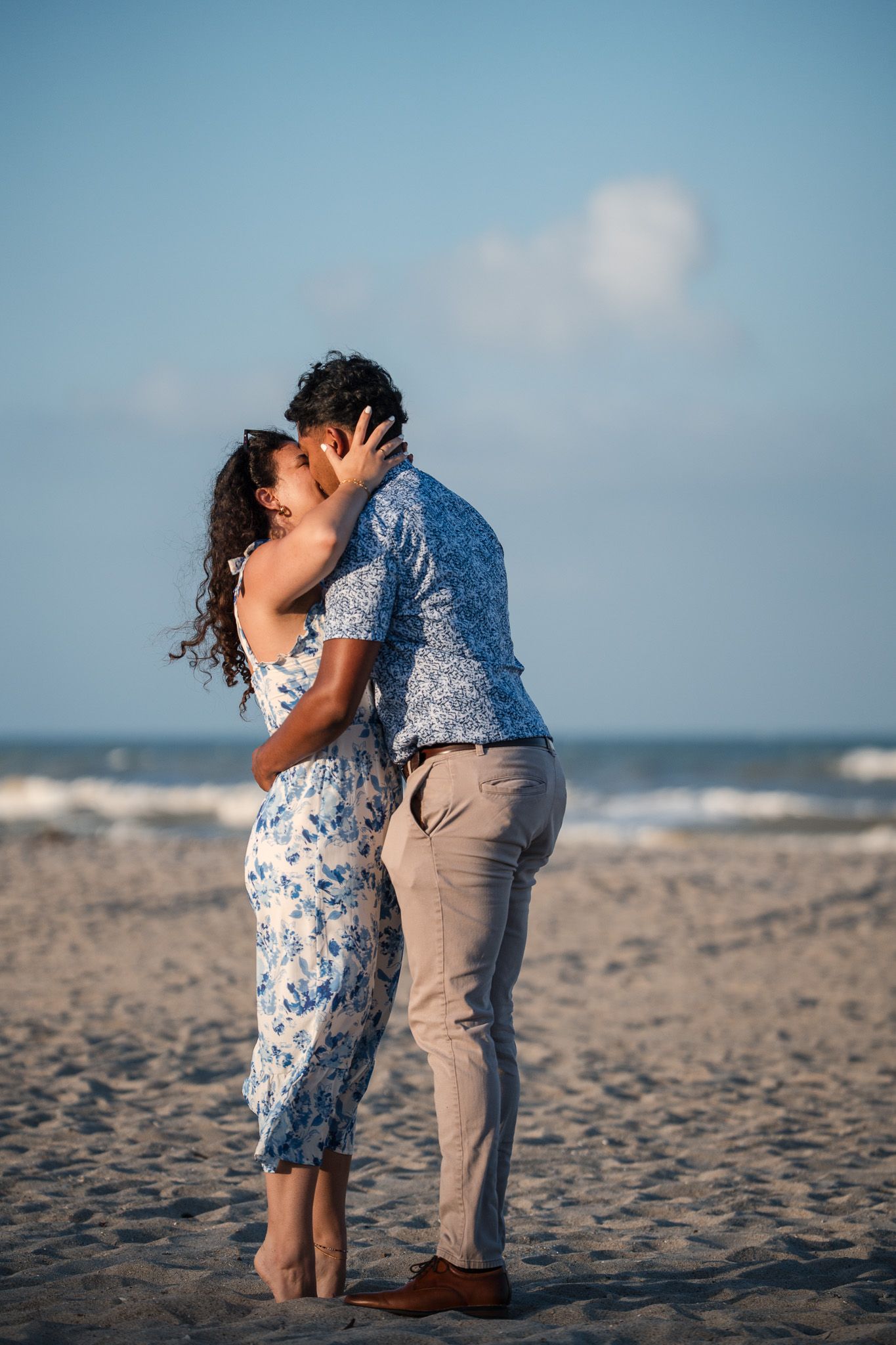 Romantic couple sharing a kiss on a sandy beach at sunset, woman in floral dress and man in blue shirt.