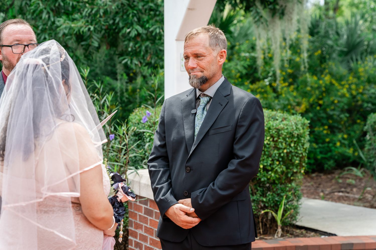 A man in a dark suit watches emotionally as a bride in a white veil stands at an outdoor wedding ceremony.