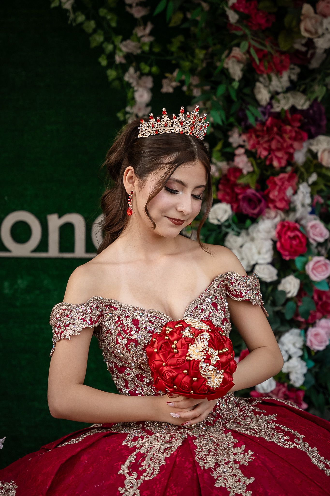 Young woman in red gold quinceañera gown with crown holds red bouquet before floral backdrop.