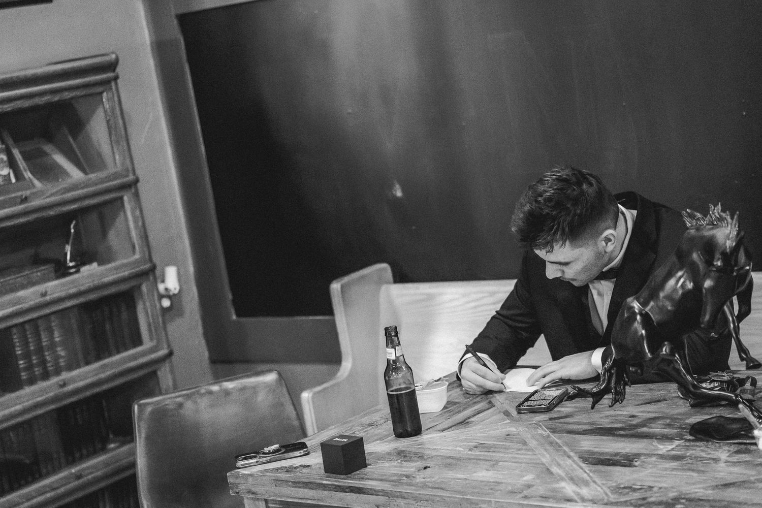 Groom in black suit writes wedding vows at a rustic wooden table in black and white.