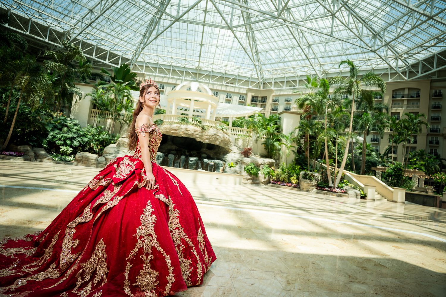 Woman in red gold embroidered ballgown poses in elegant glass atrium with lush greenery and arched architecture.