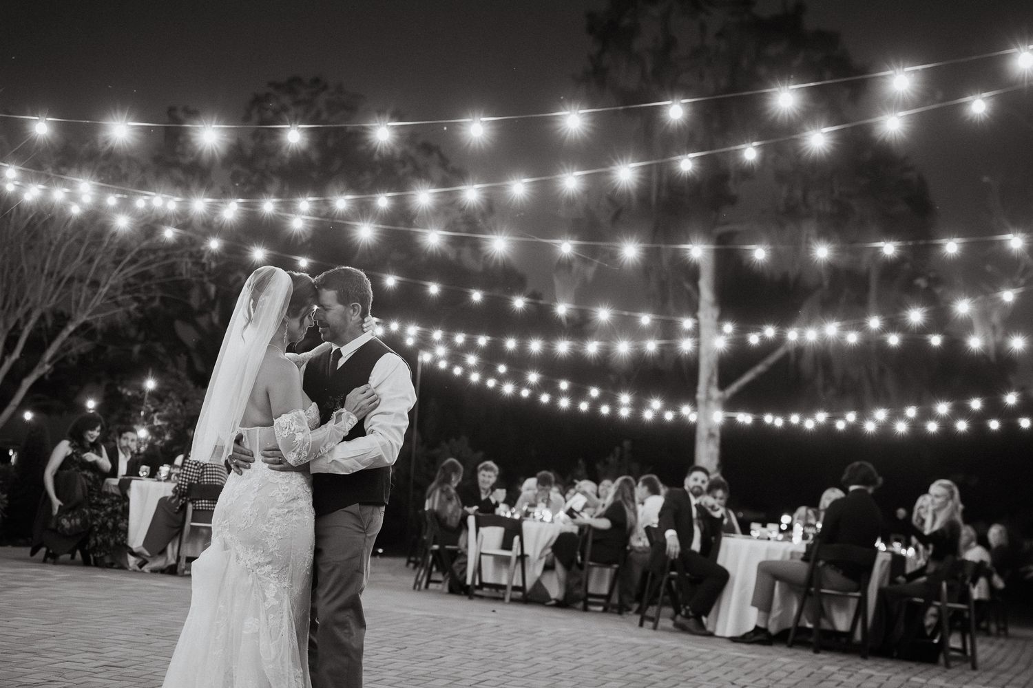 Bride and groom share first dance under string lights at elegant outdoor evening wedding reception.