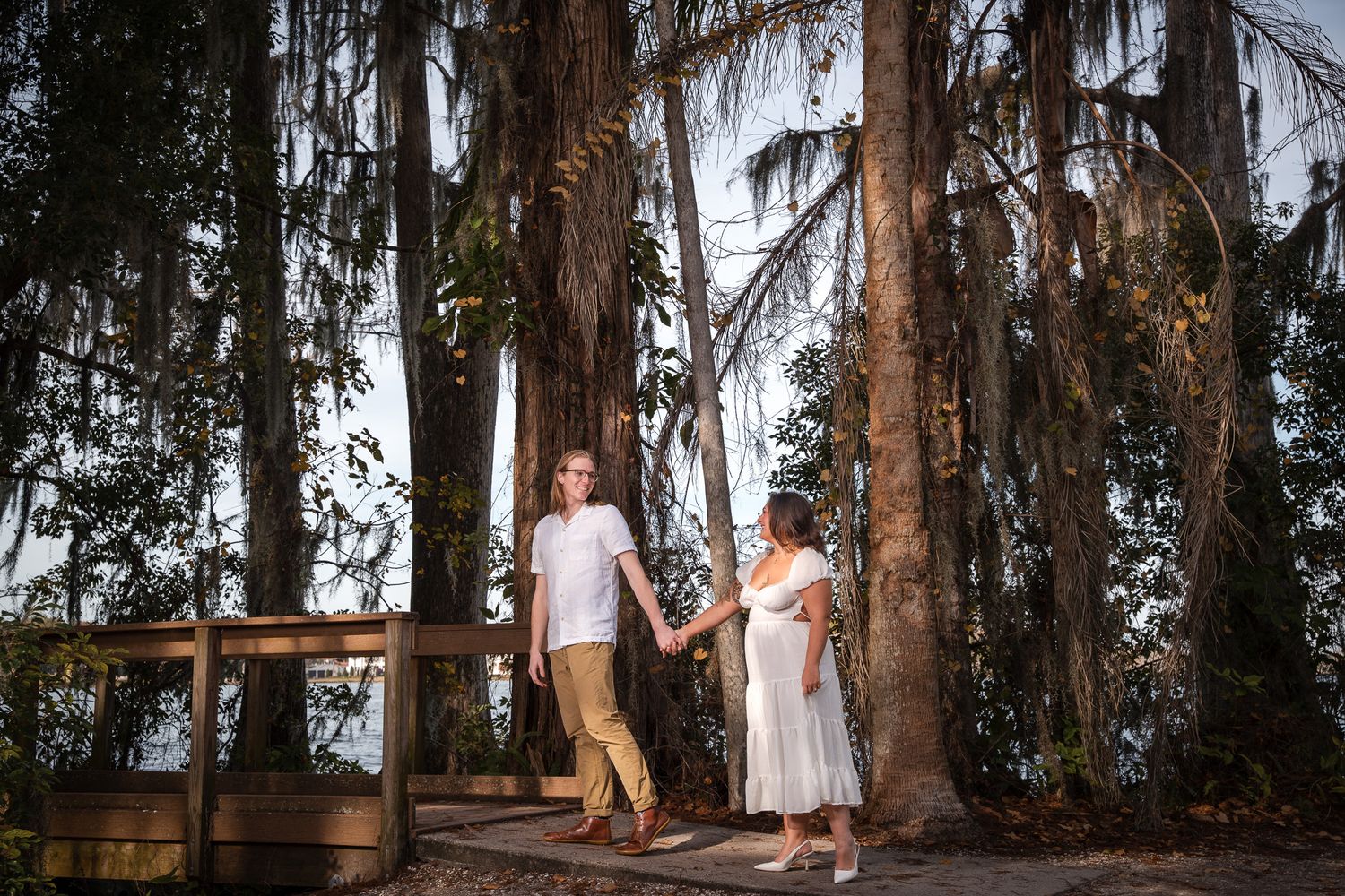 Couple holding hands on a wooden deck surrounded by Spanish moss-draped trees at dusk.