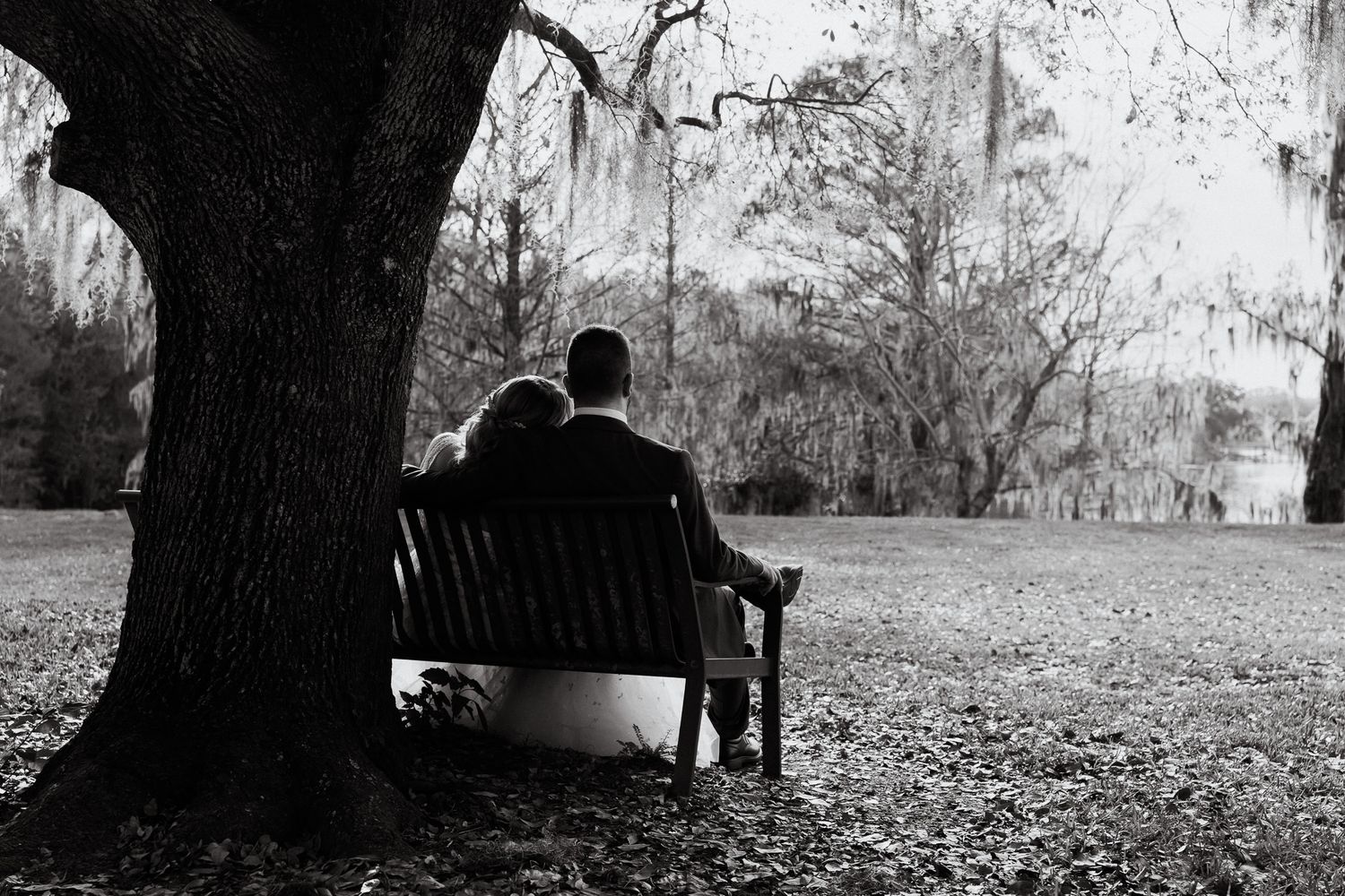 Black and white photo of bride and groom sitting together on a bench under a large tree in a serene park setting.